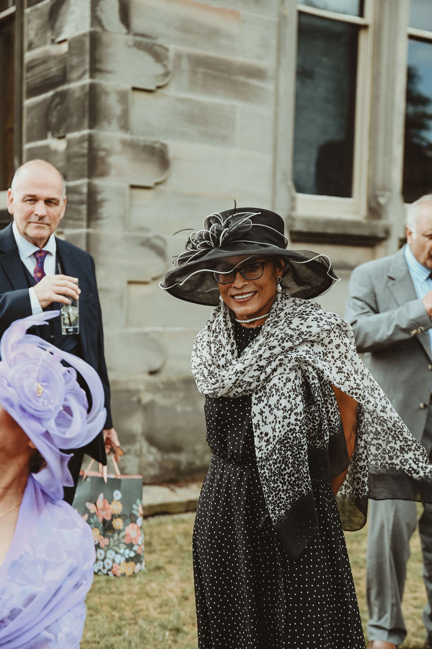 Elegantly dressed wedding guests mingle outside a historic stone building during a celebration.