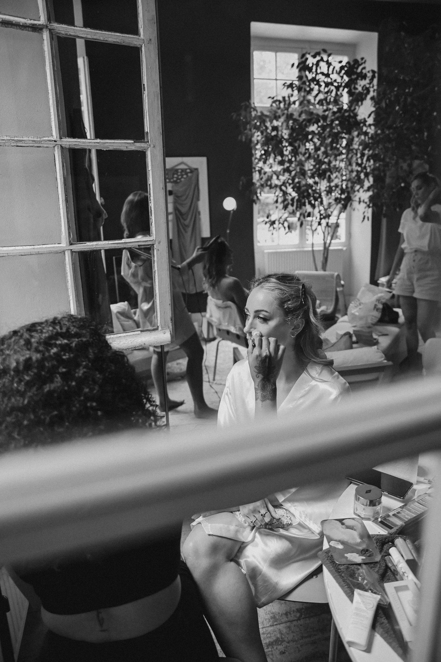 Black and white photograph showing a woman in a casual setting with a window and indoor plant in the background.