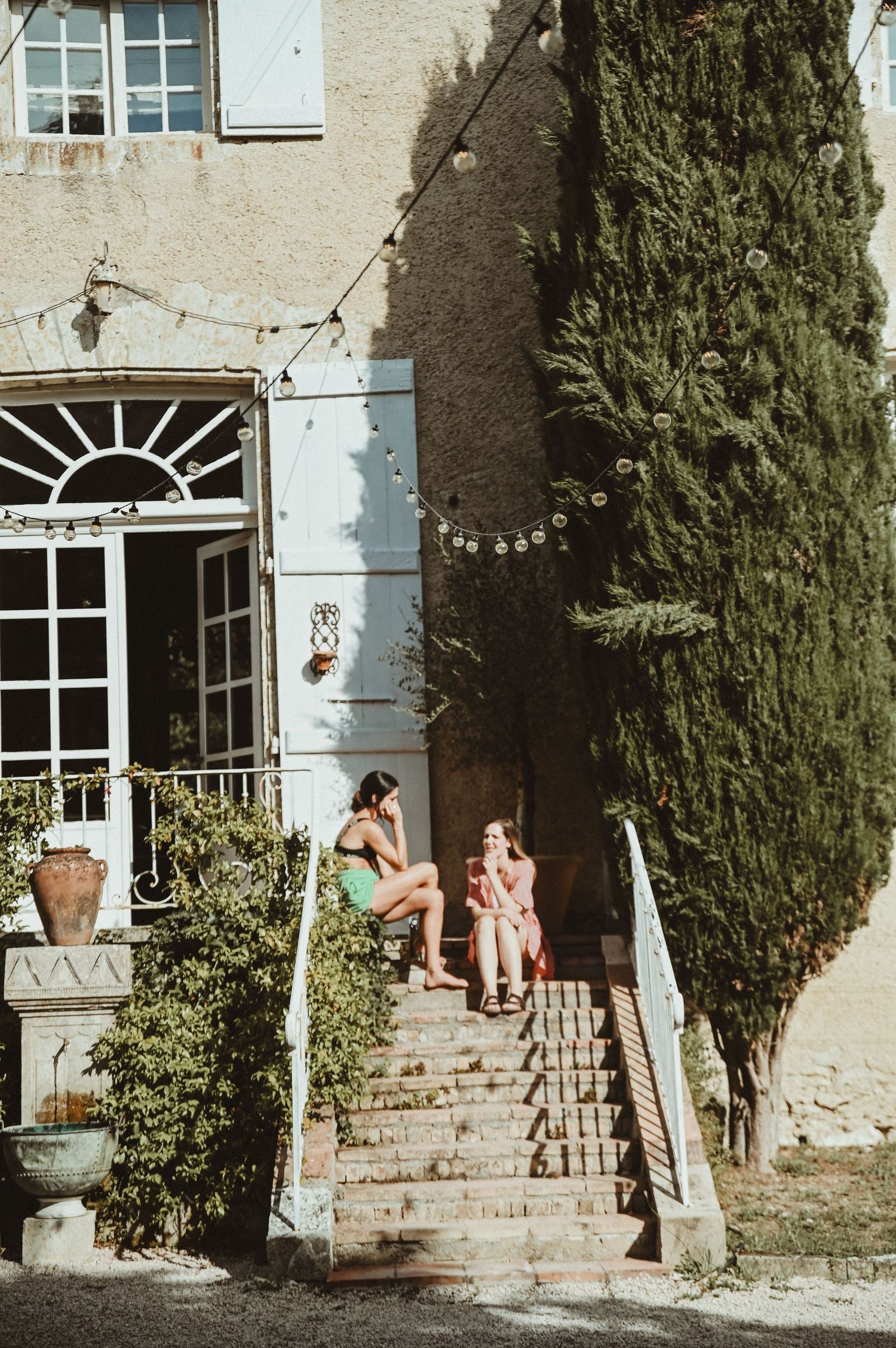 Historic stone house exterior with steps and tall cypress tree casting shadows on a sunny afternoon.