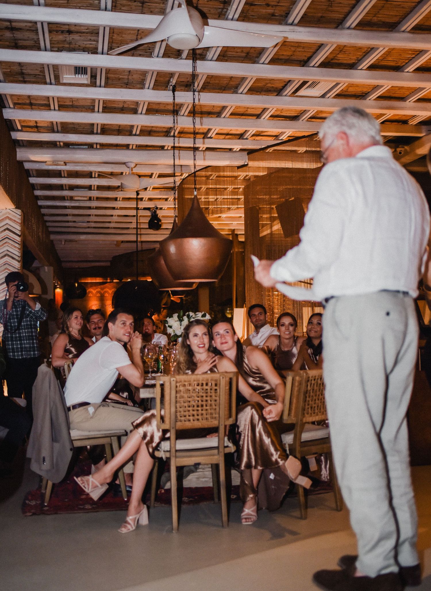 Guests enjoy a lively gathering in a rustic venue with wooden ceiling beams and copper pendant lights.