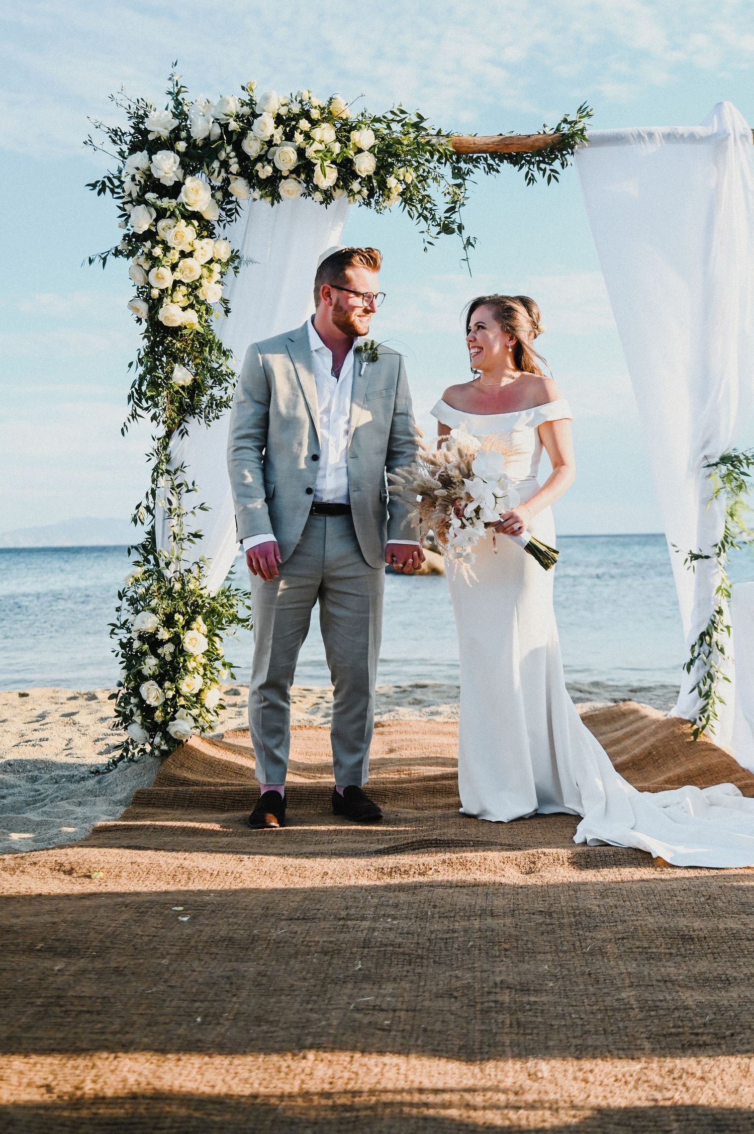 Outdoor beach wedding ceremony with couple standing under floral arch with white roses and greenery at sunset.