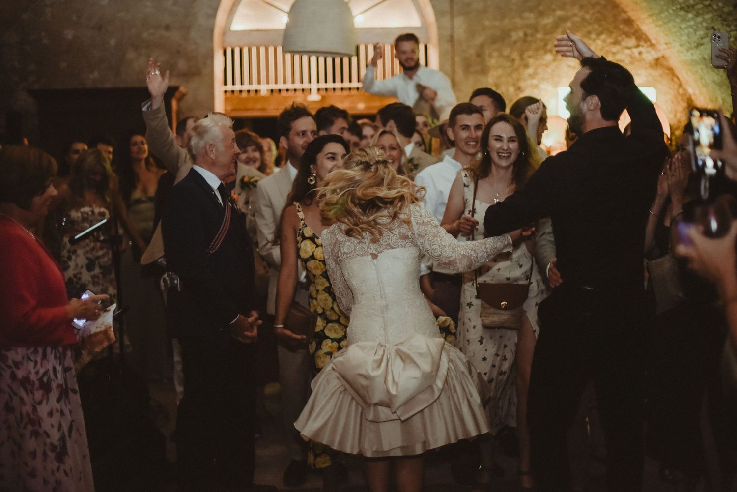 A joyous wedding celebration with guests dancing in a historic stone venue with vaulted ceilings.