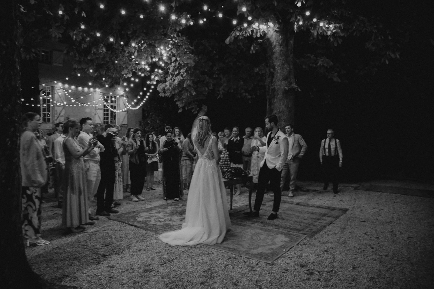 A wedding ceremony outdoors at night with string lights illuminating guests gathered on a decorated outdoor space.