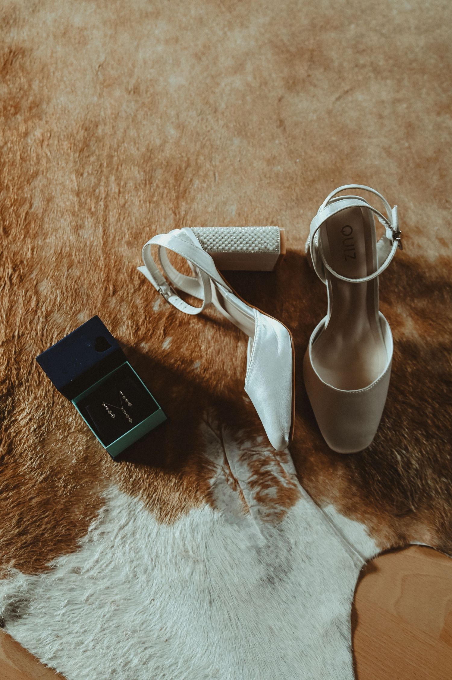 Wedding accessories including white shoes and ring box arranged on furry rug with moody lighting.