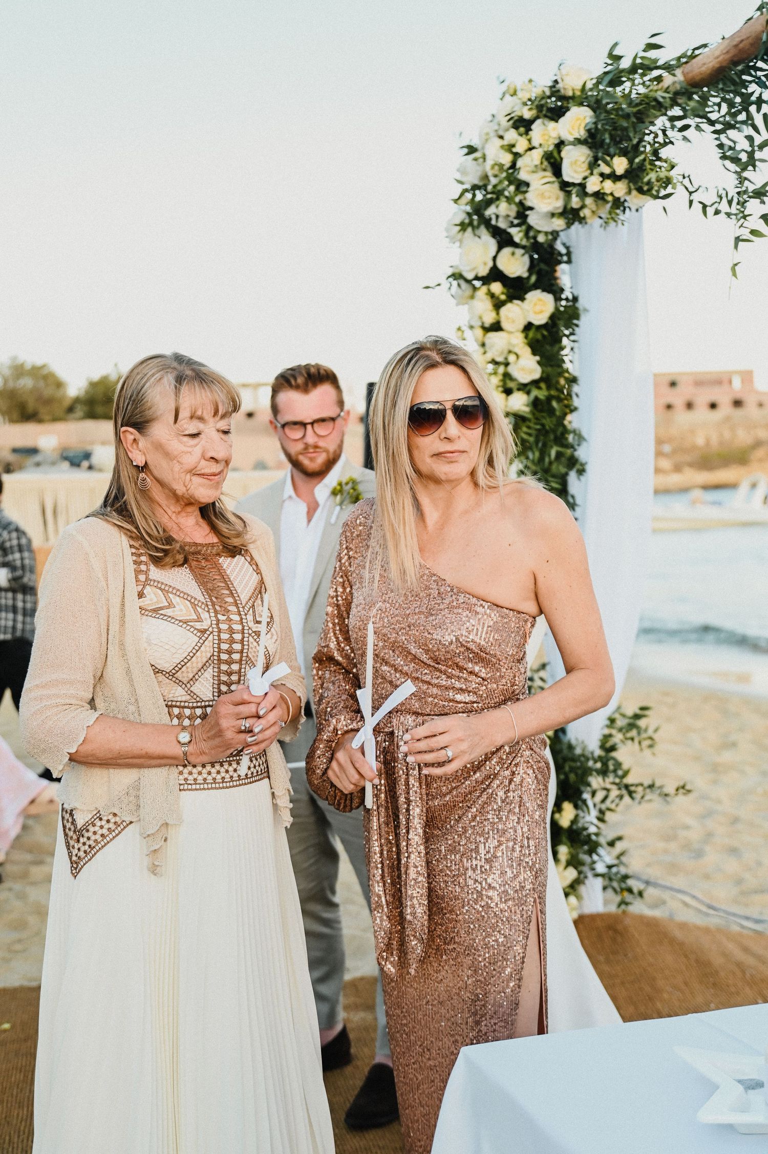 Wedding guests in elegant attire gather beneath floral arch at beach celebration.