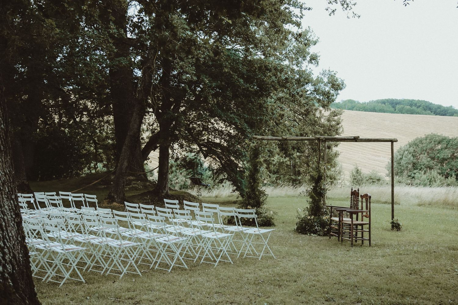 Outdoor wedding ceremony setup with white folding chairs and wooden arch in a serene wooded setting.