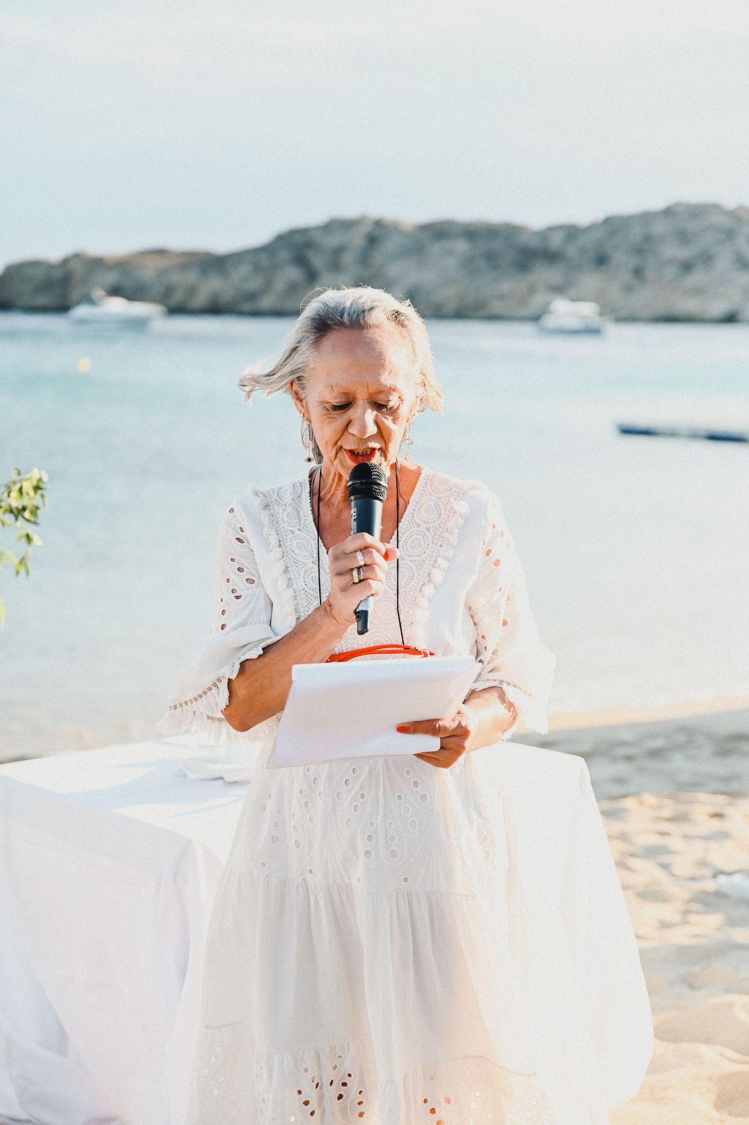 Wedding officiant in white lace dress holds ceremony papers on sunny beach with ocean views.