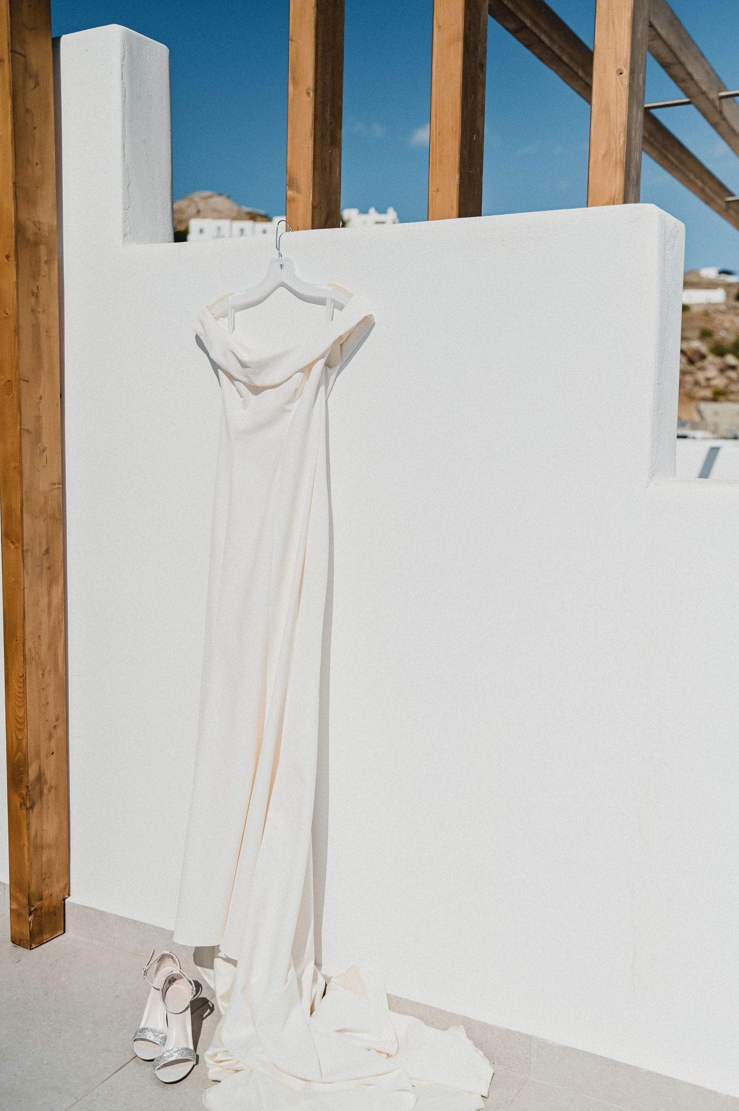 Flowing white wedding dress hanging on white wall with bridal shoes beneath.