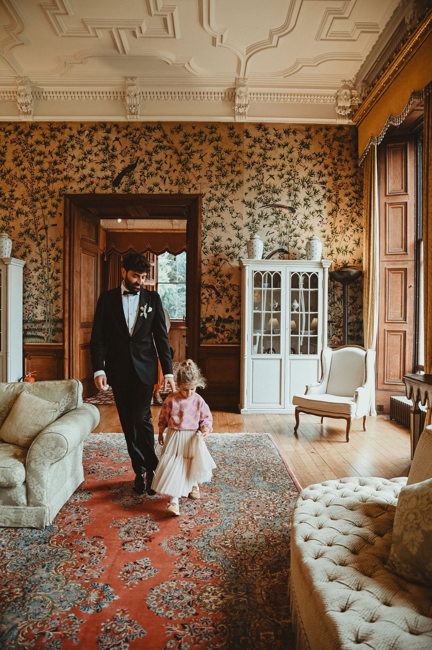 Elegant room with floral wallpaper and ornate ceiling featuring tufted furniture and wooden doorways.