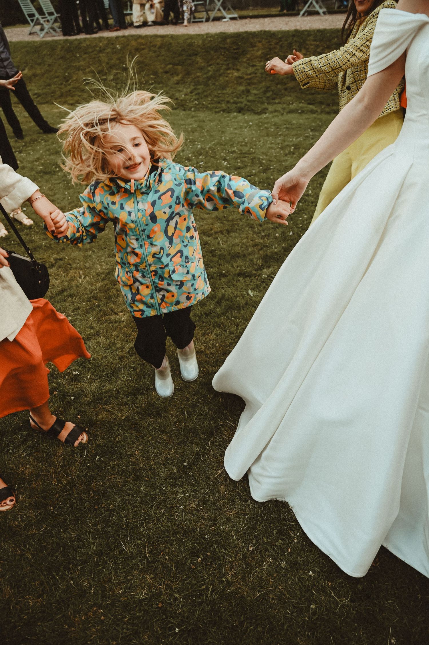 A child in a colorful patterned shirt jumps with joy while dancing on grass near a wedding dress.