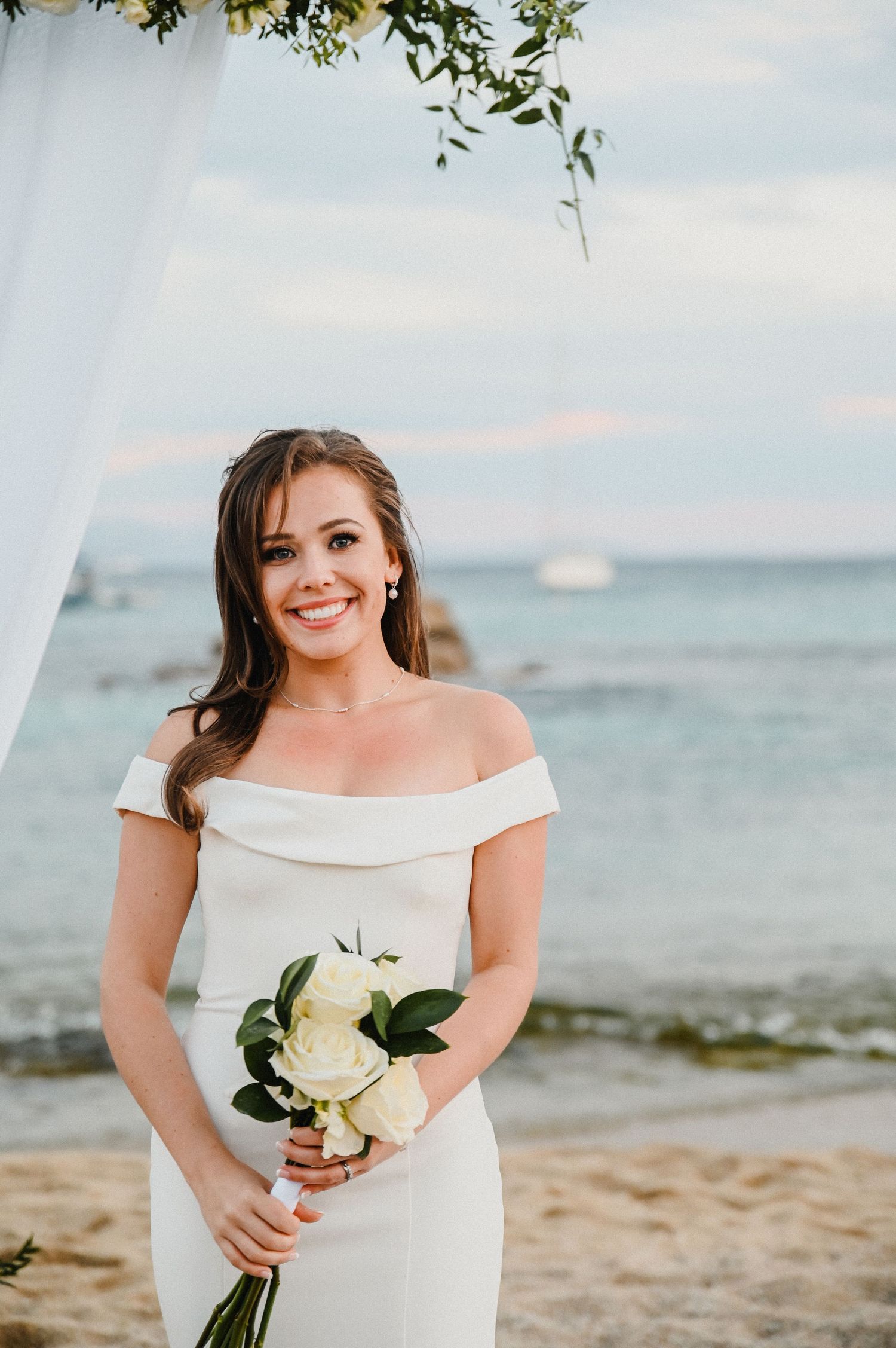 Beach wedding portrait under white floral arch with ocean and boats in background.