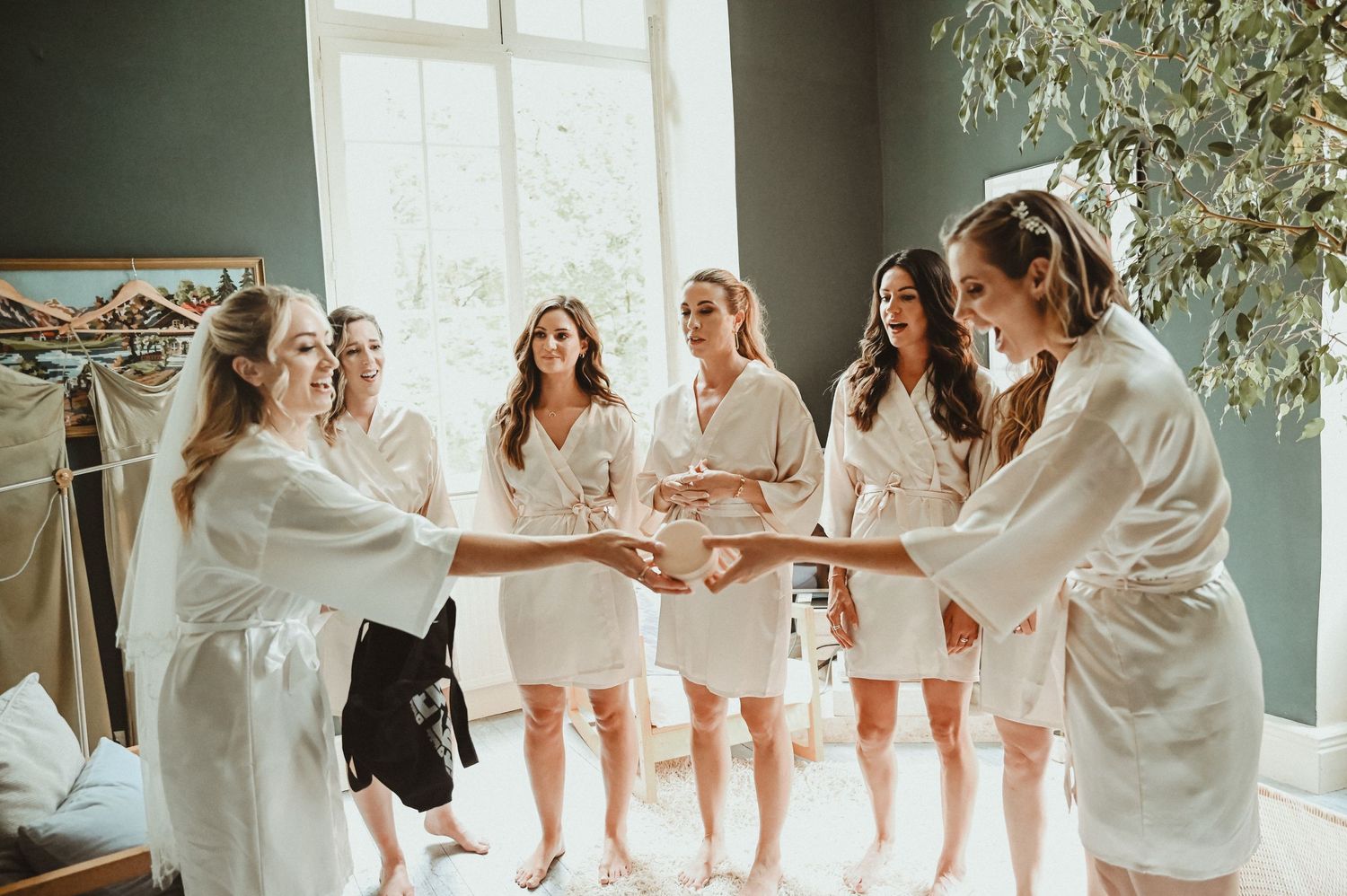Group in white robes celebrates during wedding morning preparations in sunlit room.