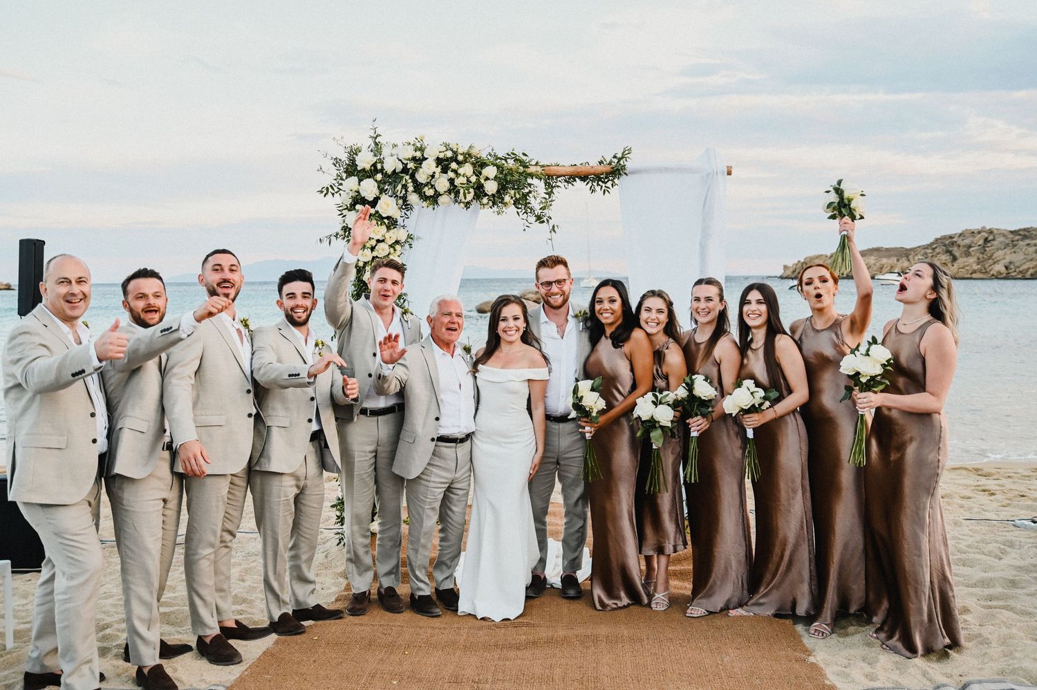 Wedding party group photo on beach platform with floral arch and ocean backdrop.