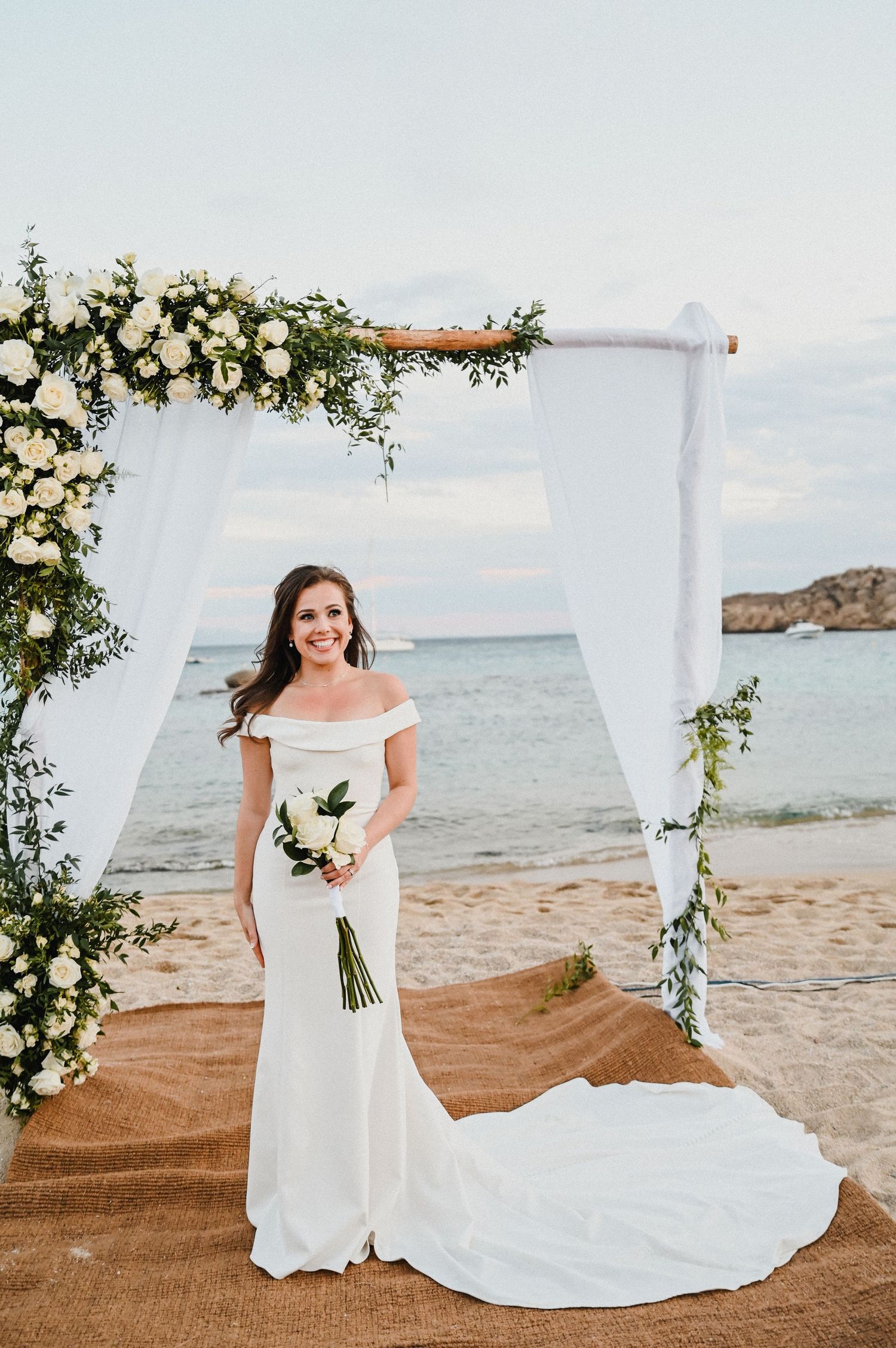 Elegant beach wedding ceremony setup with white floral arch and flowing curtains against an ocean backdrop.