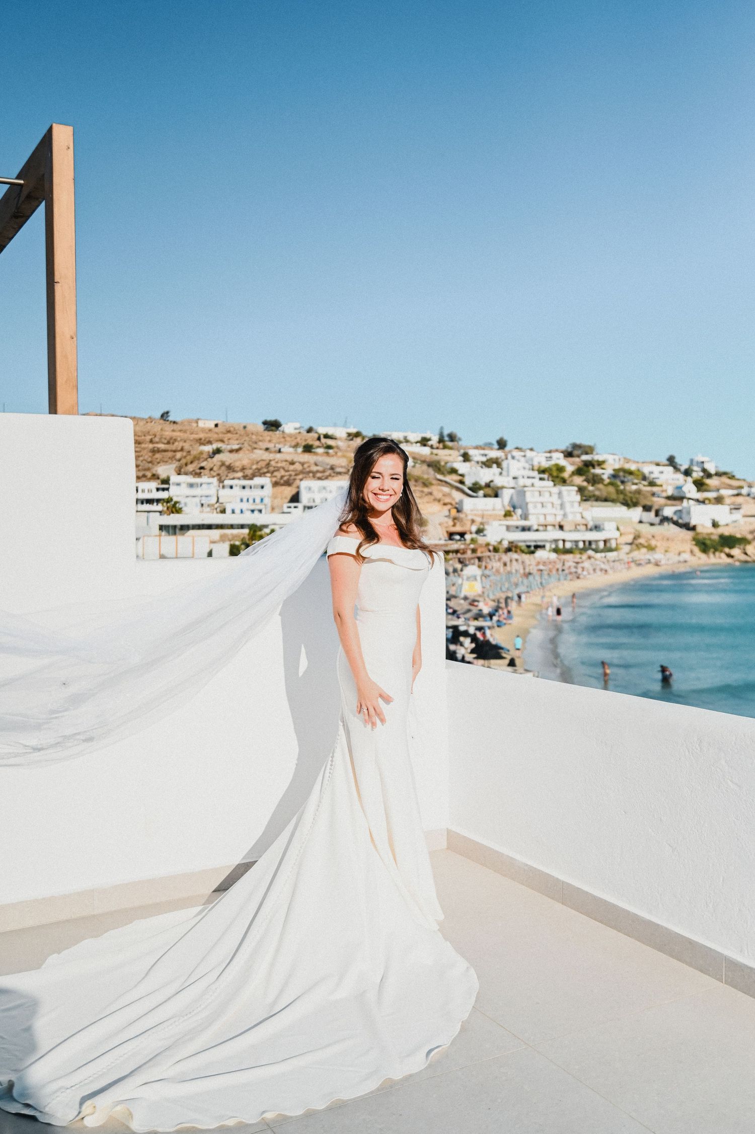 Bride in flowing white gown poses on terrace overlooking Mediterranean beach town.