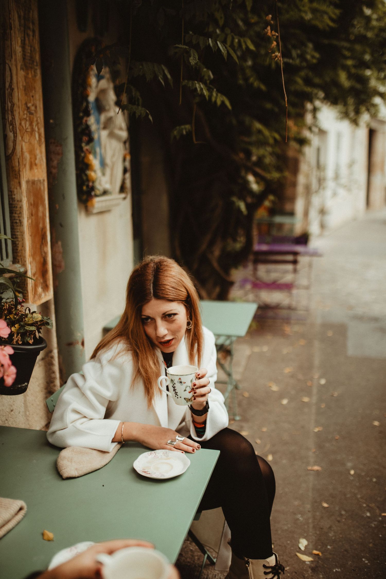 A person sits at an outdoor cafe table holding a coffee cup on a cozy autumn day.