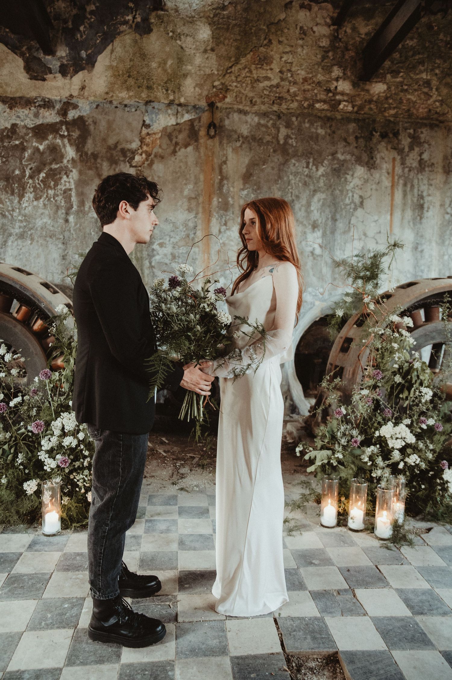 A couple stands facing each other during their wedding ceremony surrounded by candlelight and floral arrangements.