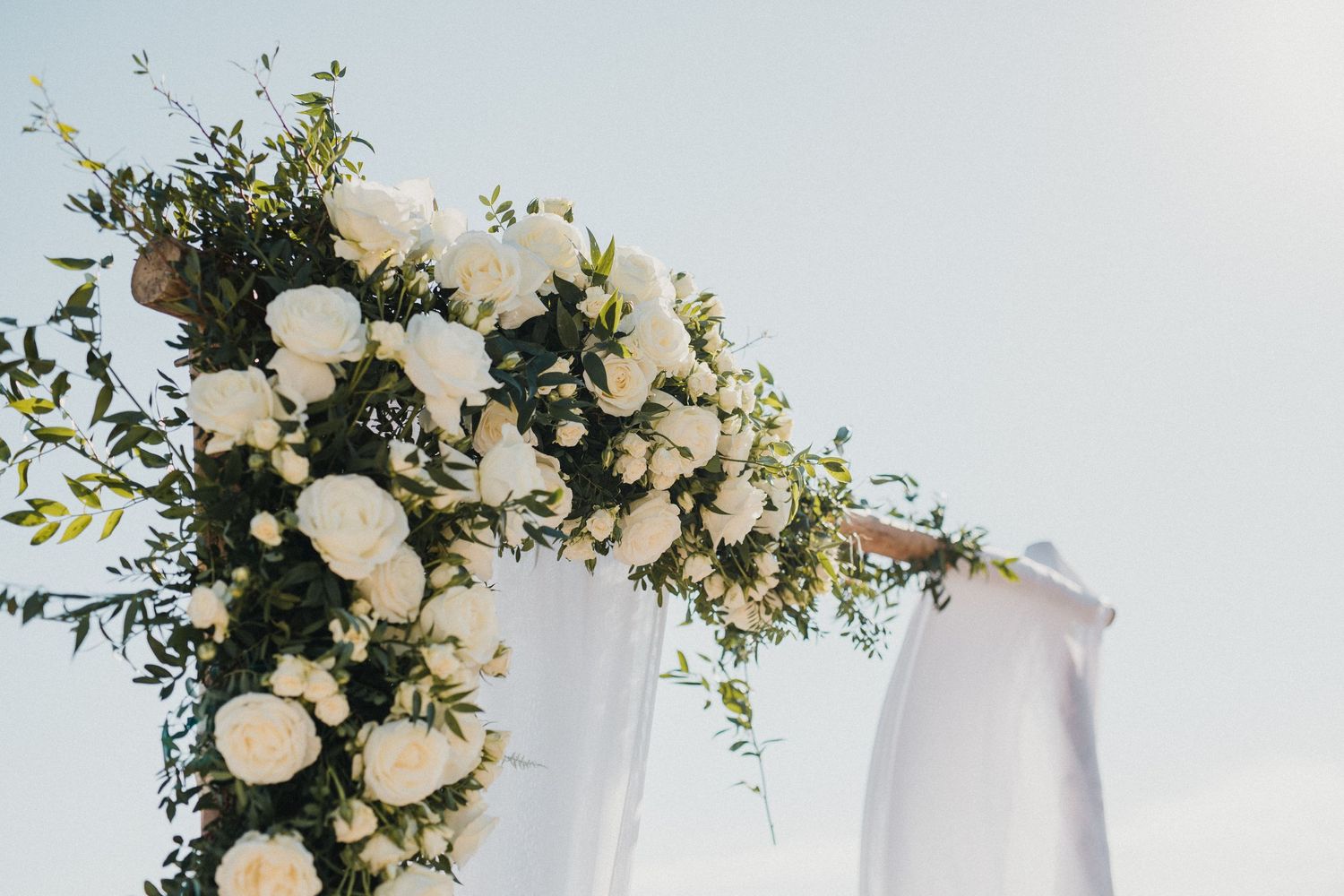 White floral arch with roses and greenery decorates an outdoor wedding ceremony space.