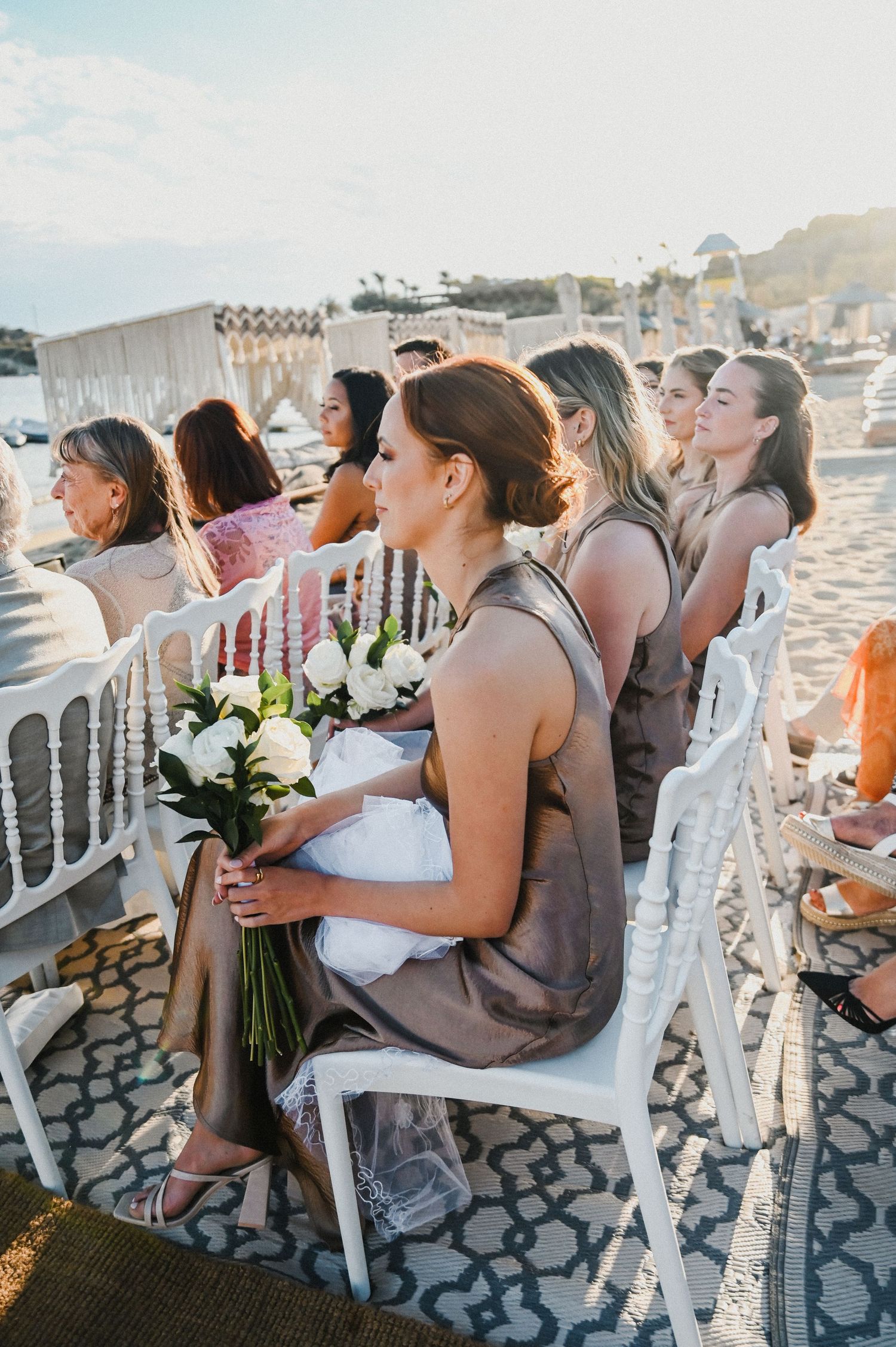 Wedding guests seated in white chairs holding bouquets during beachside ceremony.