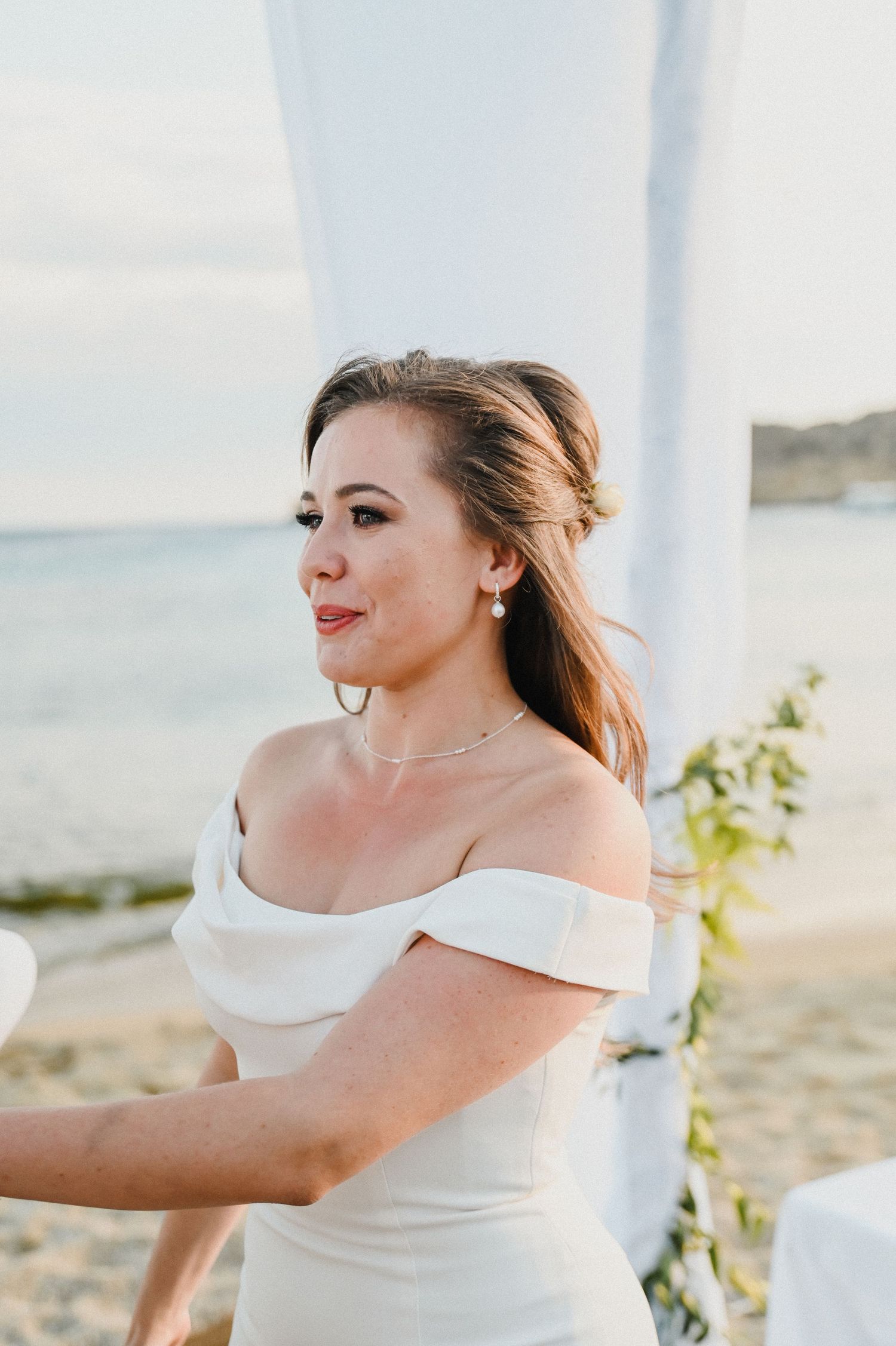A bride in an off-shoulder white wedding dress poses against a seaside backdrop during golden hour.