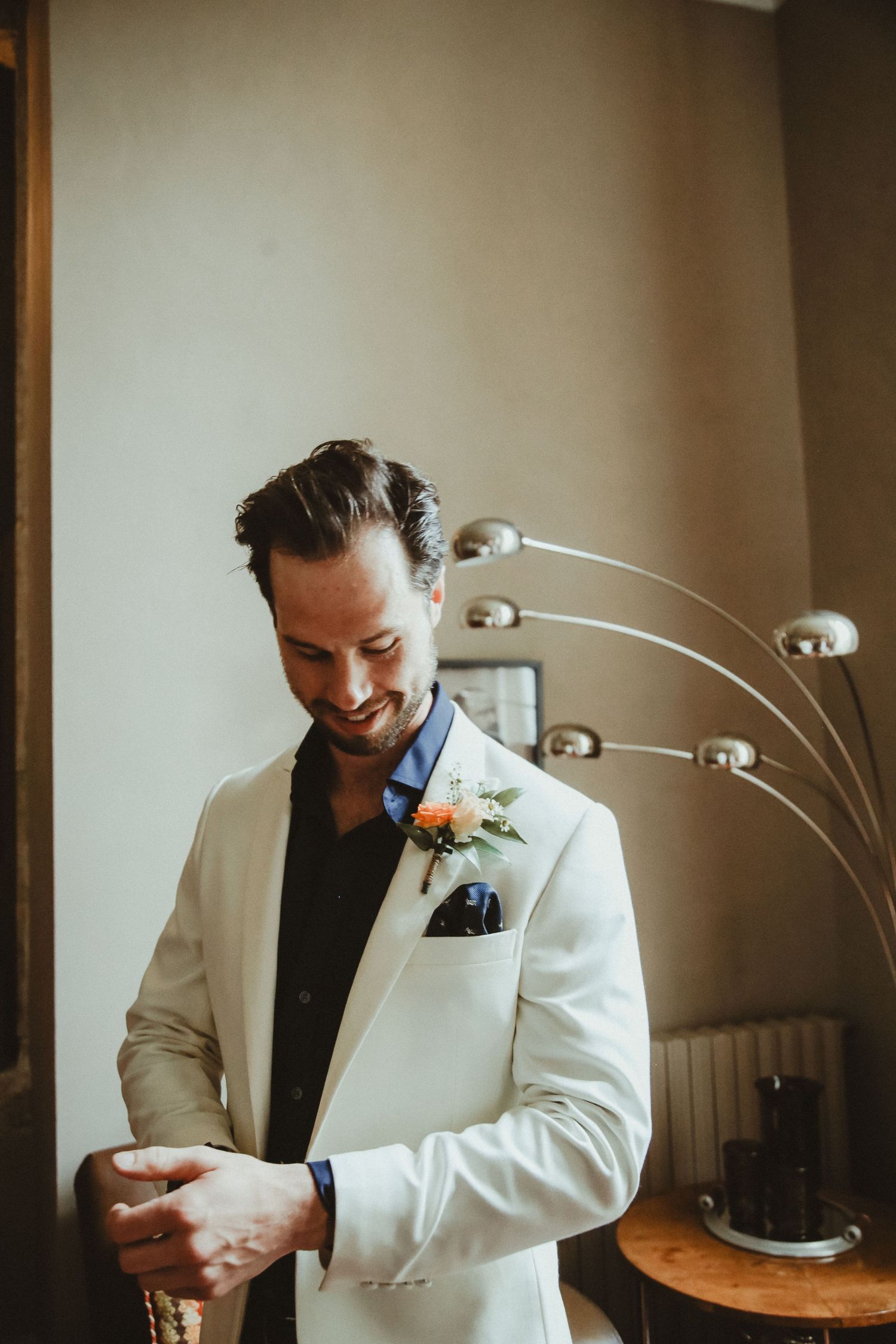 A groom in a white tuxedo jacket with boutonniere adjusts his cuff.
