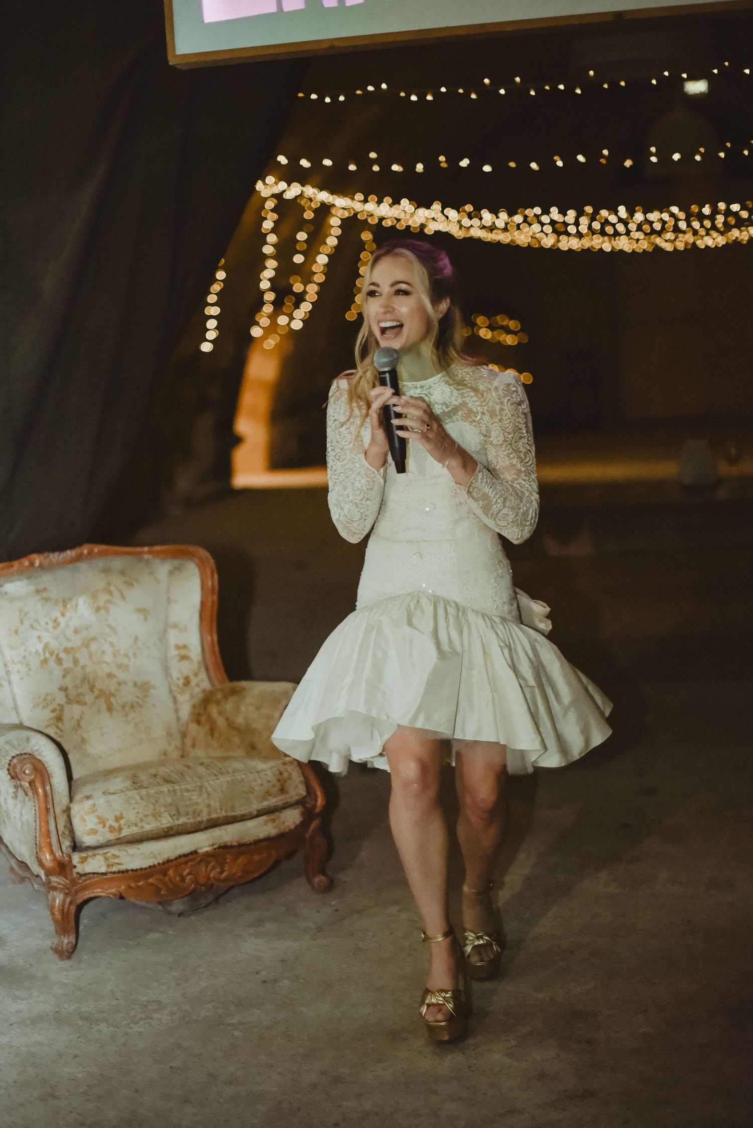 A person in a short white lace dress performs with a microphone under twinkling string lights next to a vintage couch.