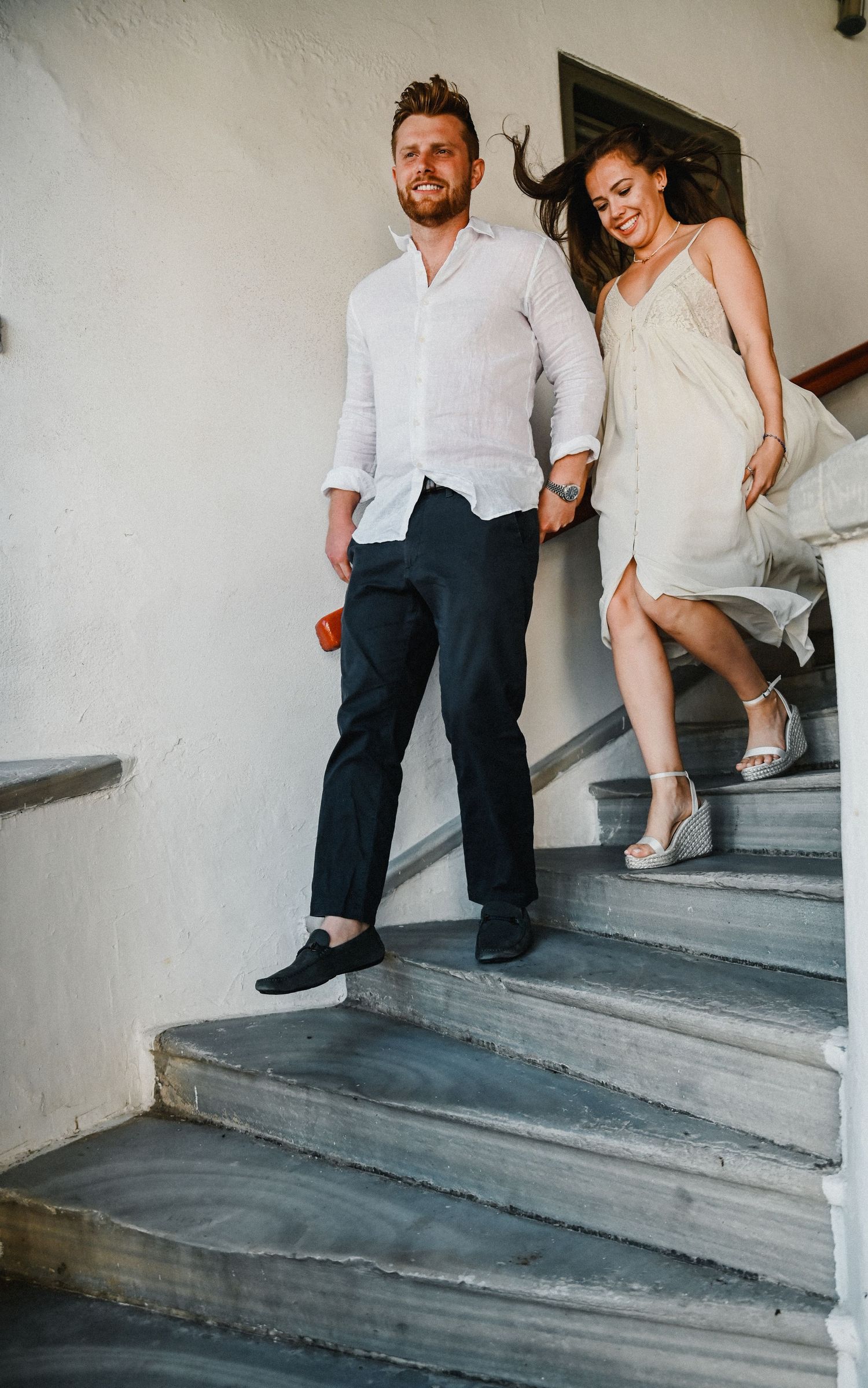 Couple in casual wedding attire descending white stone steps together in a romantic Mediterranean setting.