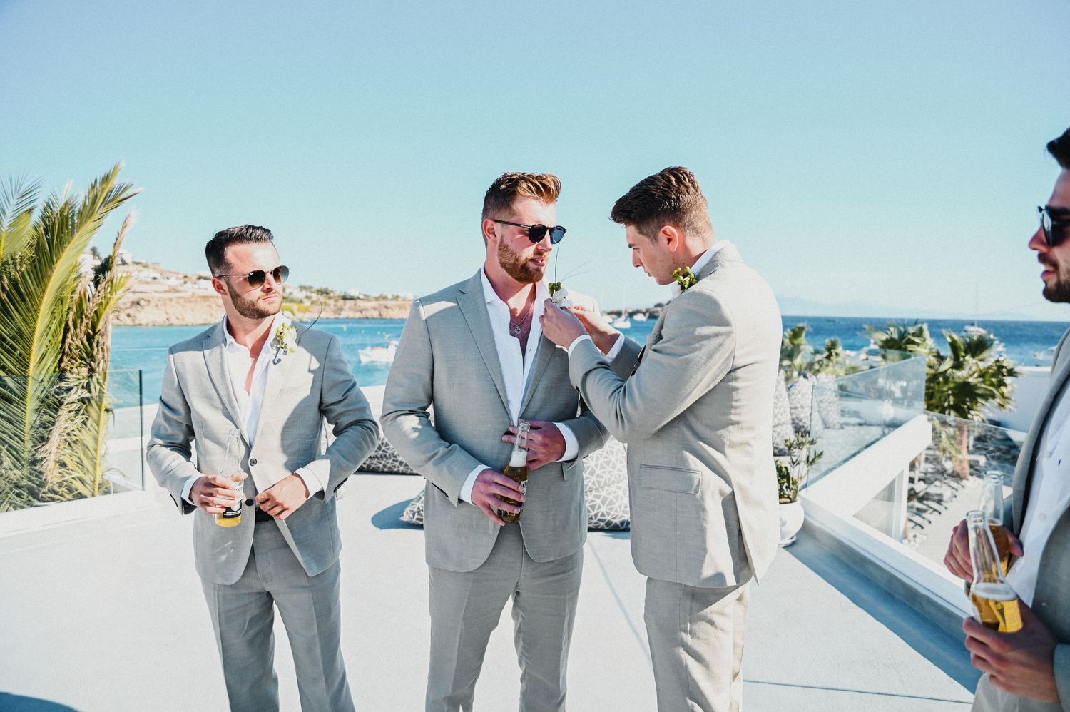 Groomsmen in light gray suits toast with drinks on a scenic white balcony overlooking the ocean.
