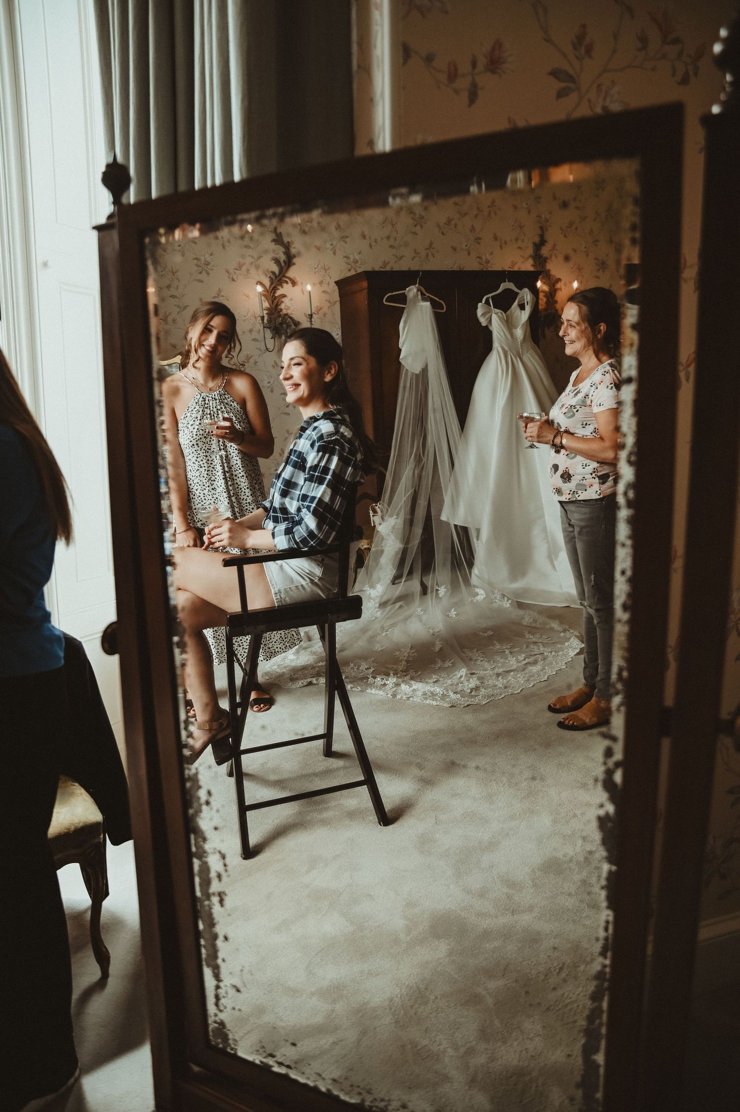 Wedding preparations reflected in an antique mirror showing white dresses hanging and friends gathering in a rustic room.