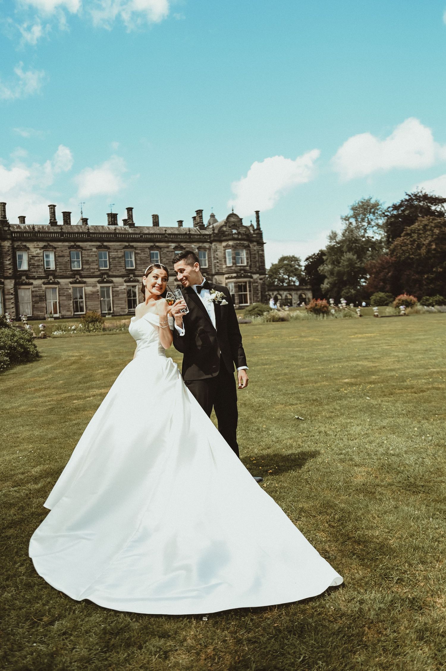 Wedding couple poses on lawn with majestic stone manor house in background.
