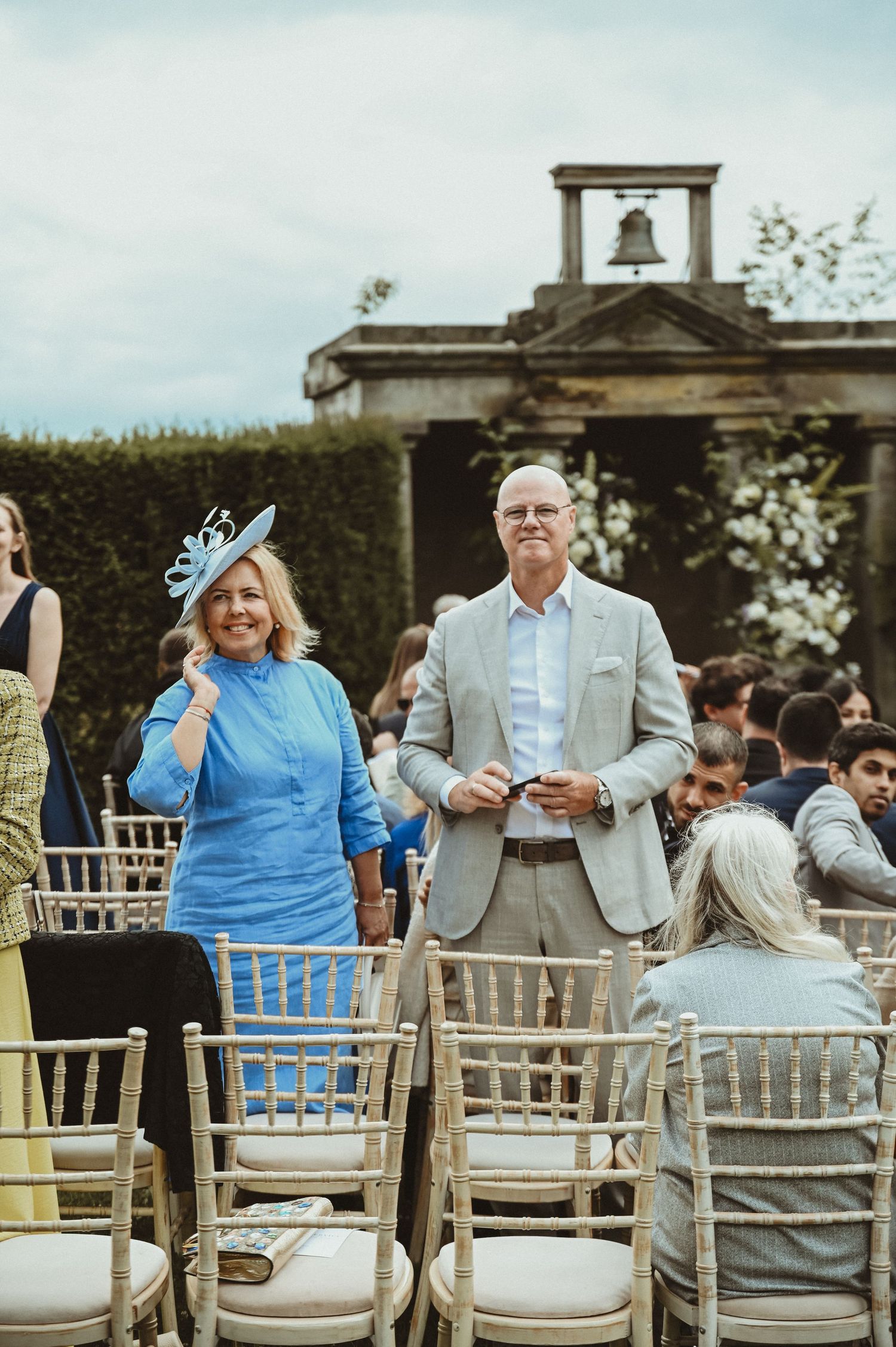 Wedding guests in formal attire stand near white chairs at an outdoor ceremony venue.
