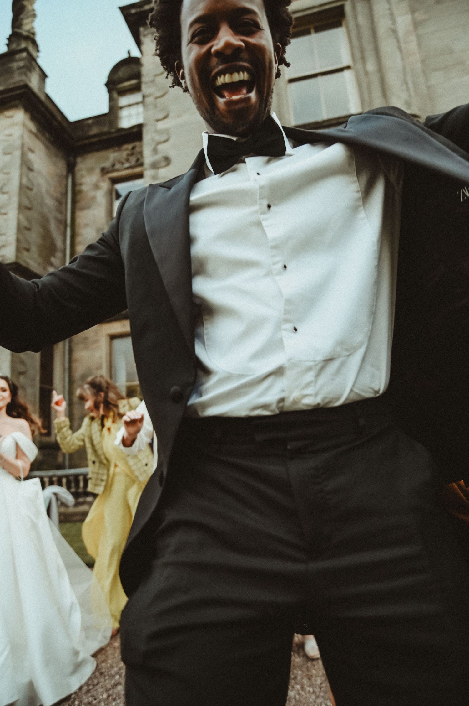 A wedding guest in formal attire dances joyfully outside a grand historic building.