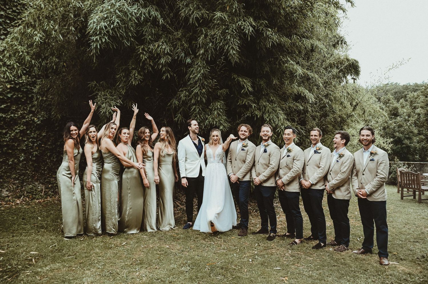 Formal wedding party lineup with bridesmaids in sage and groomsmen in tan suits outdoors.