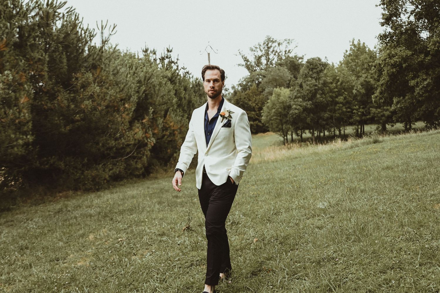 A groom in a white dinner jacket and black pants walks through a grassy field.