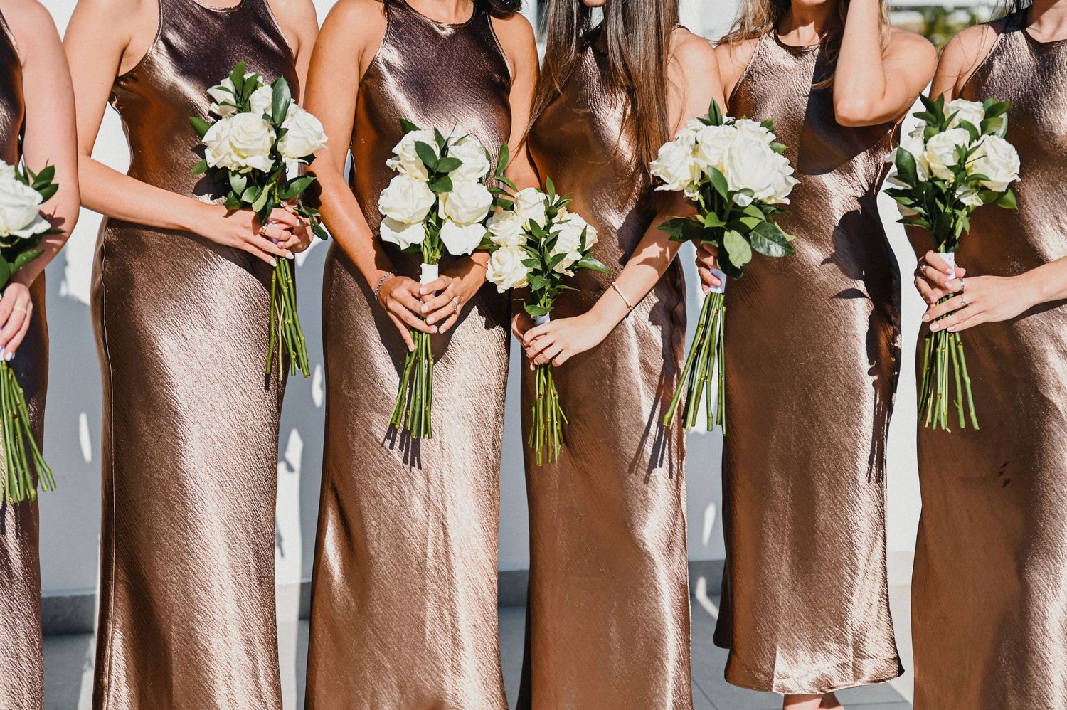 Row of bridesmaids holding white rose bouquets while wearing matching bronze satin dresses.