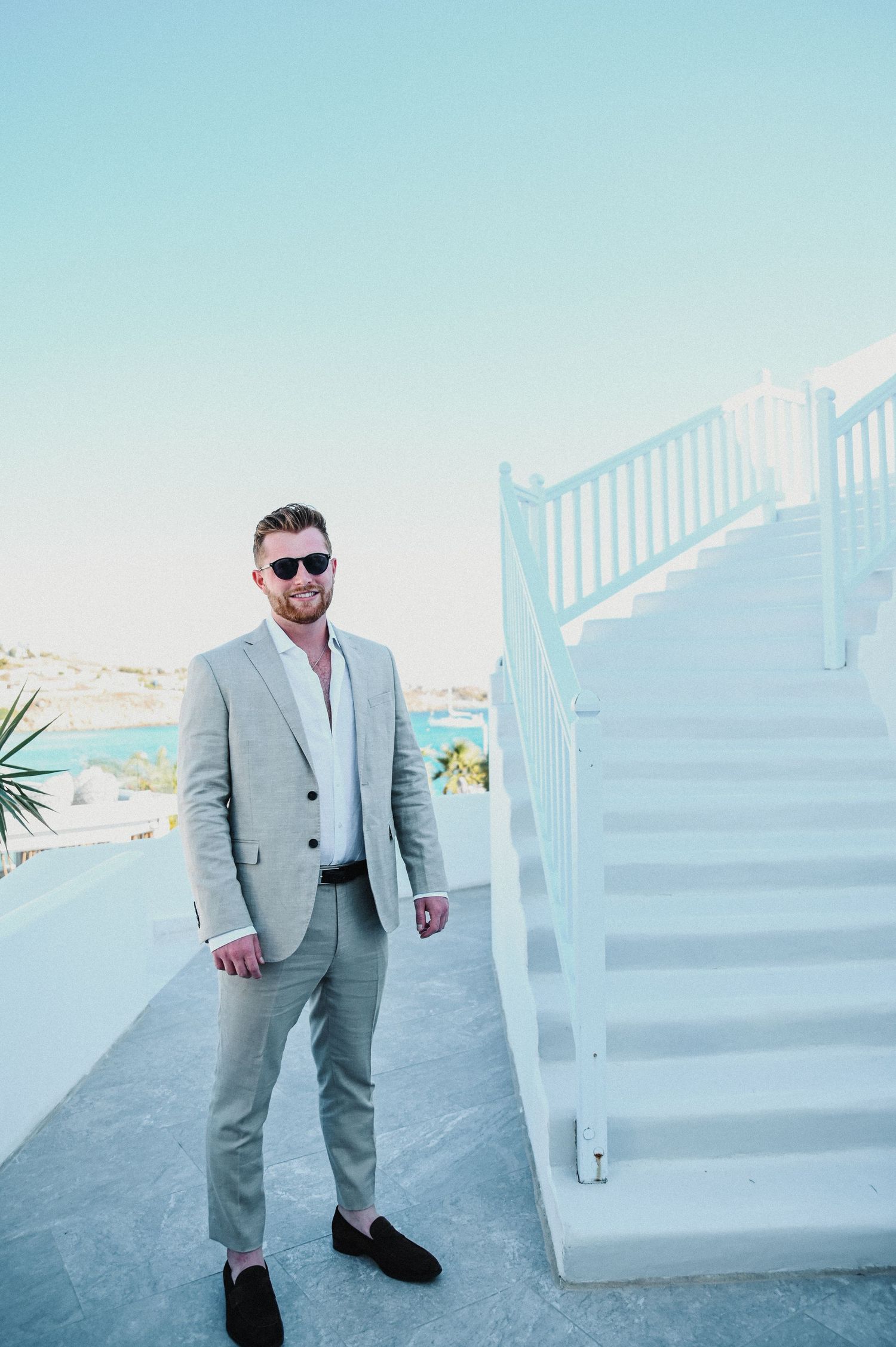 Stylish groomsman in light gray suit posing by white staircase with ocean view.