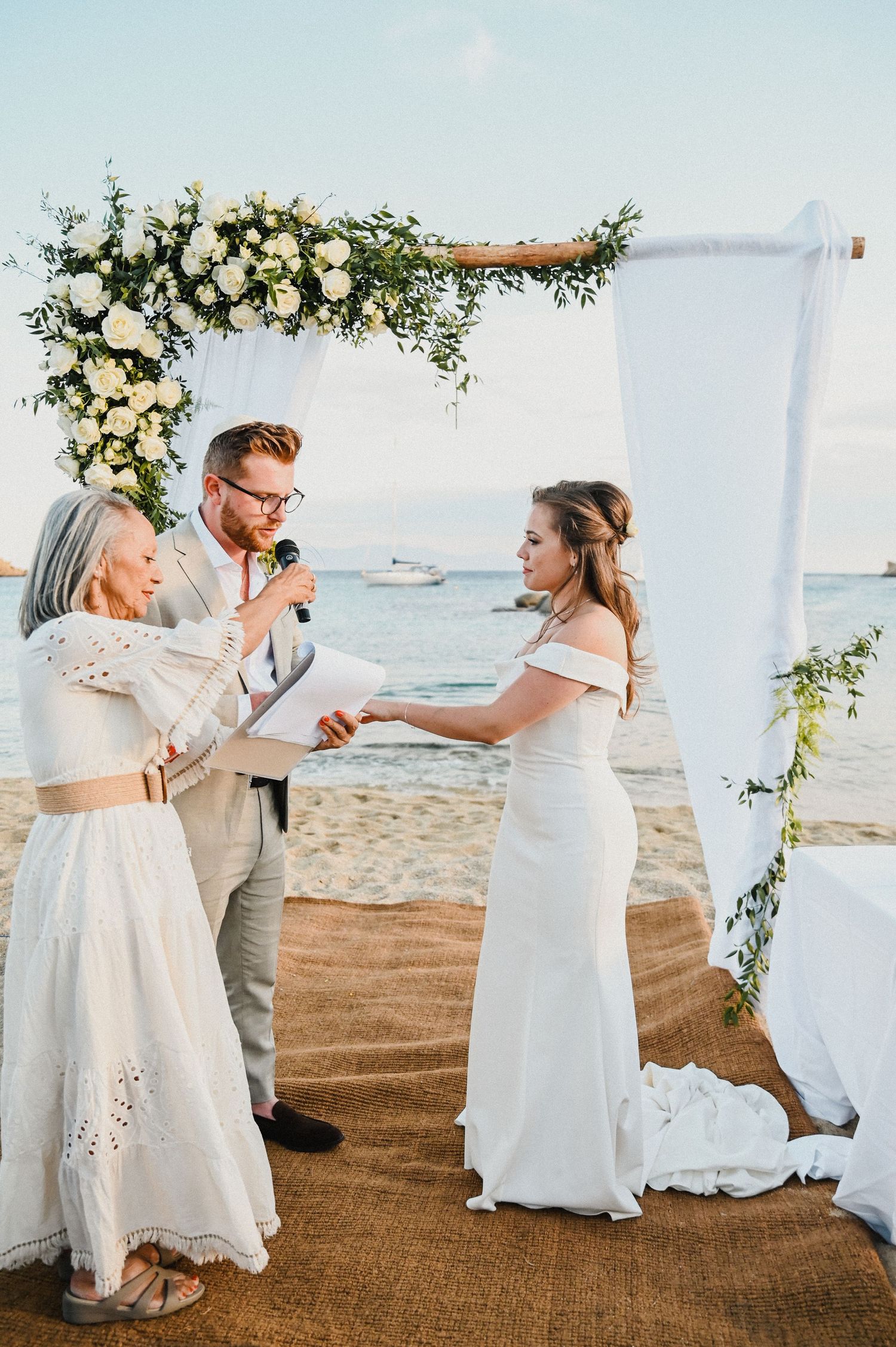Wedding ceremony on a beach with floral arch and couple exchanging vows under white draping.