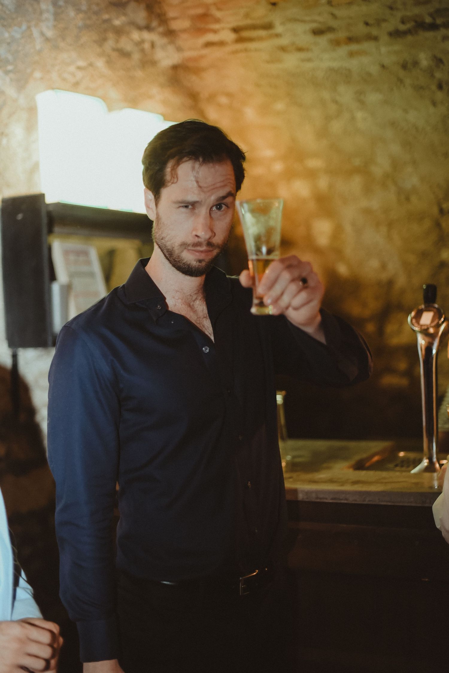 A person in a navy shirt raises a glass while standing at a bar.