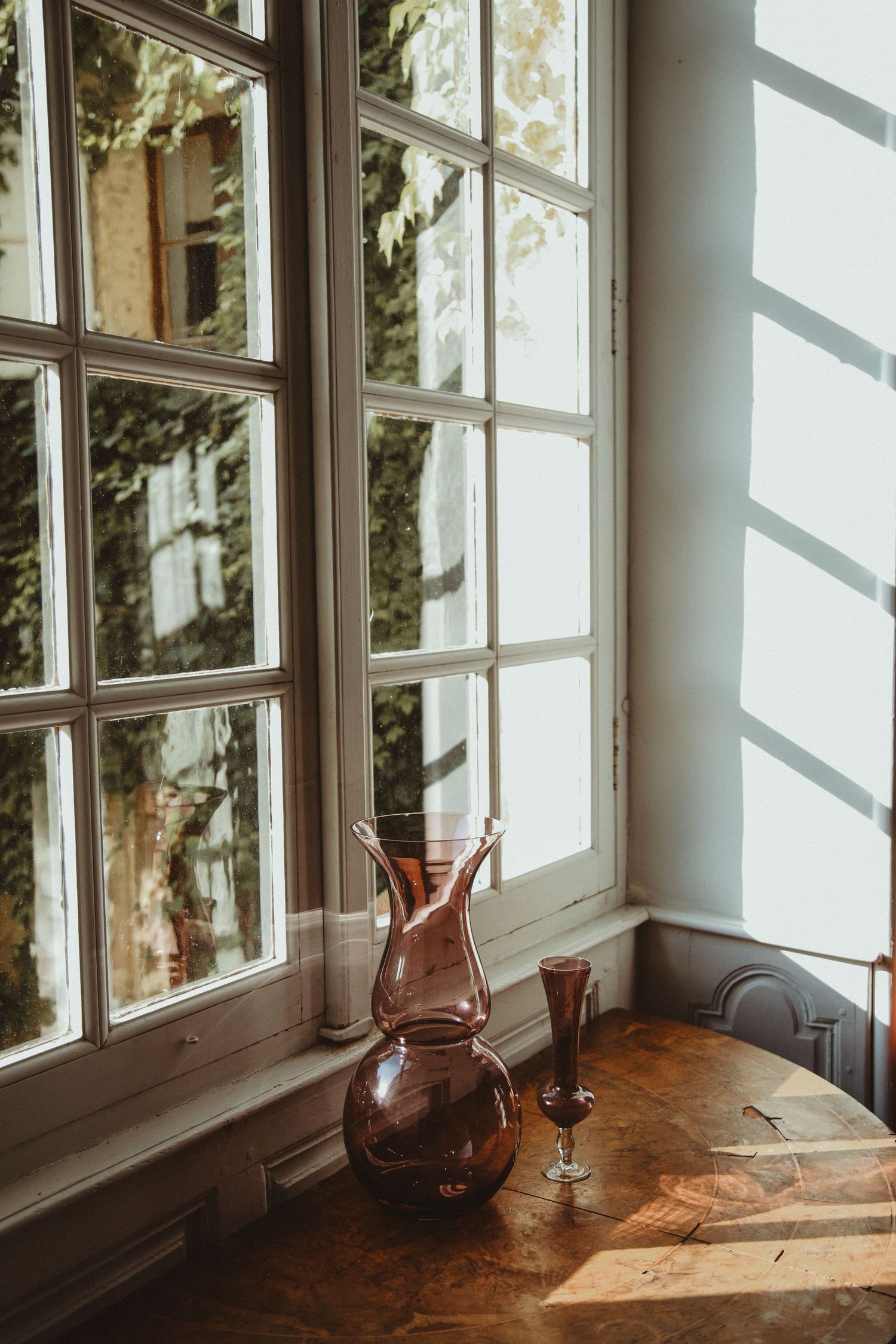 Brown glass decanter and stemware sit on a wooden table by a sunlit window casting geometric shadows.