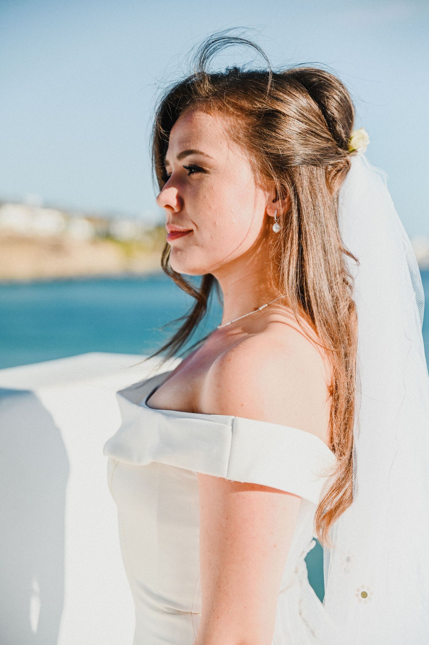 Side profile portrait of bride with flowing veil against blue ocean background.