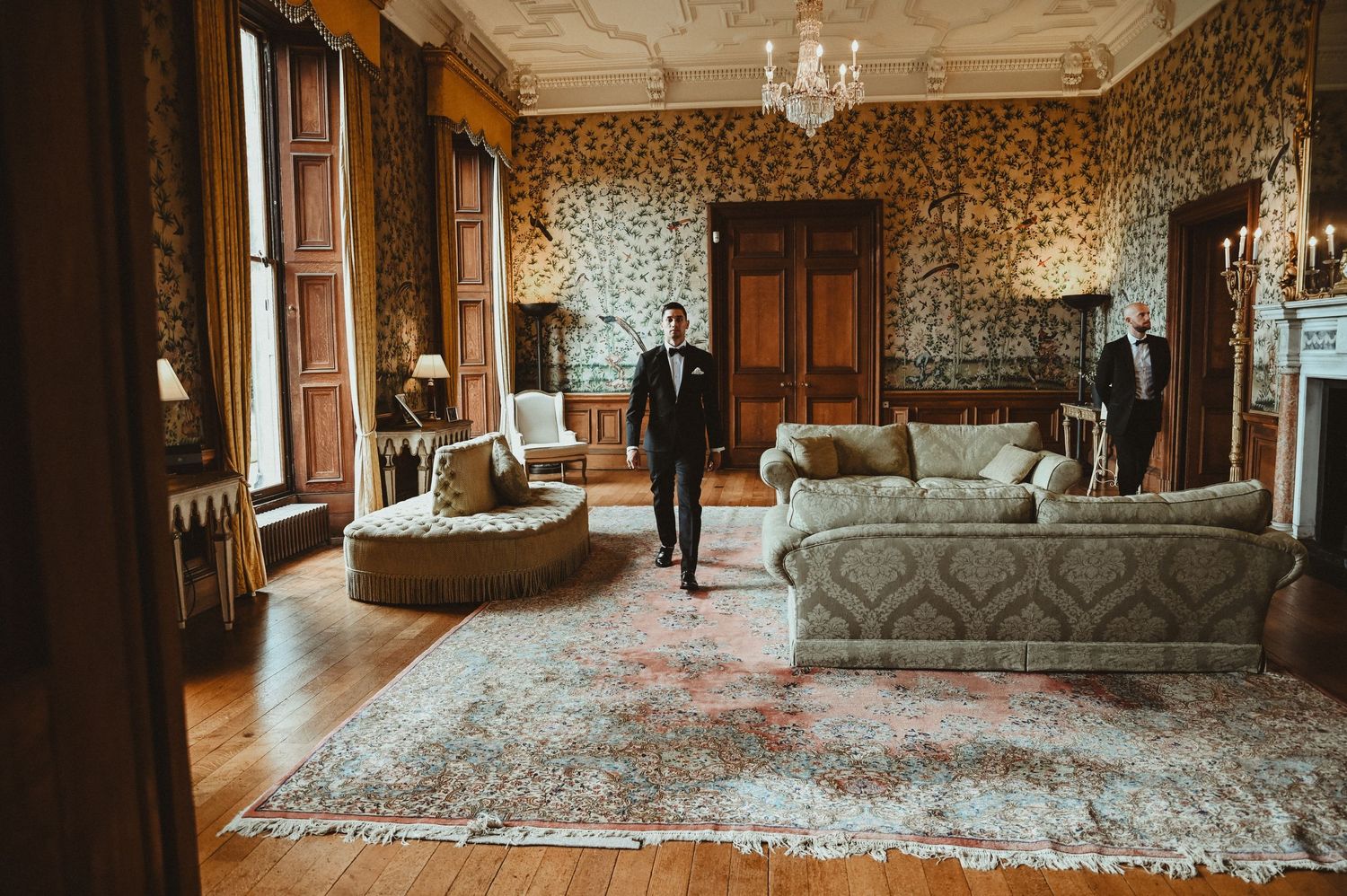 An elegant vintage room with ornate wallpaper, large windows, and period furniture.