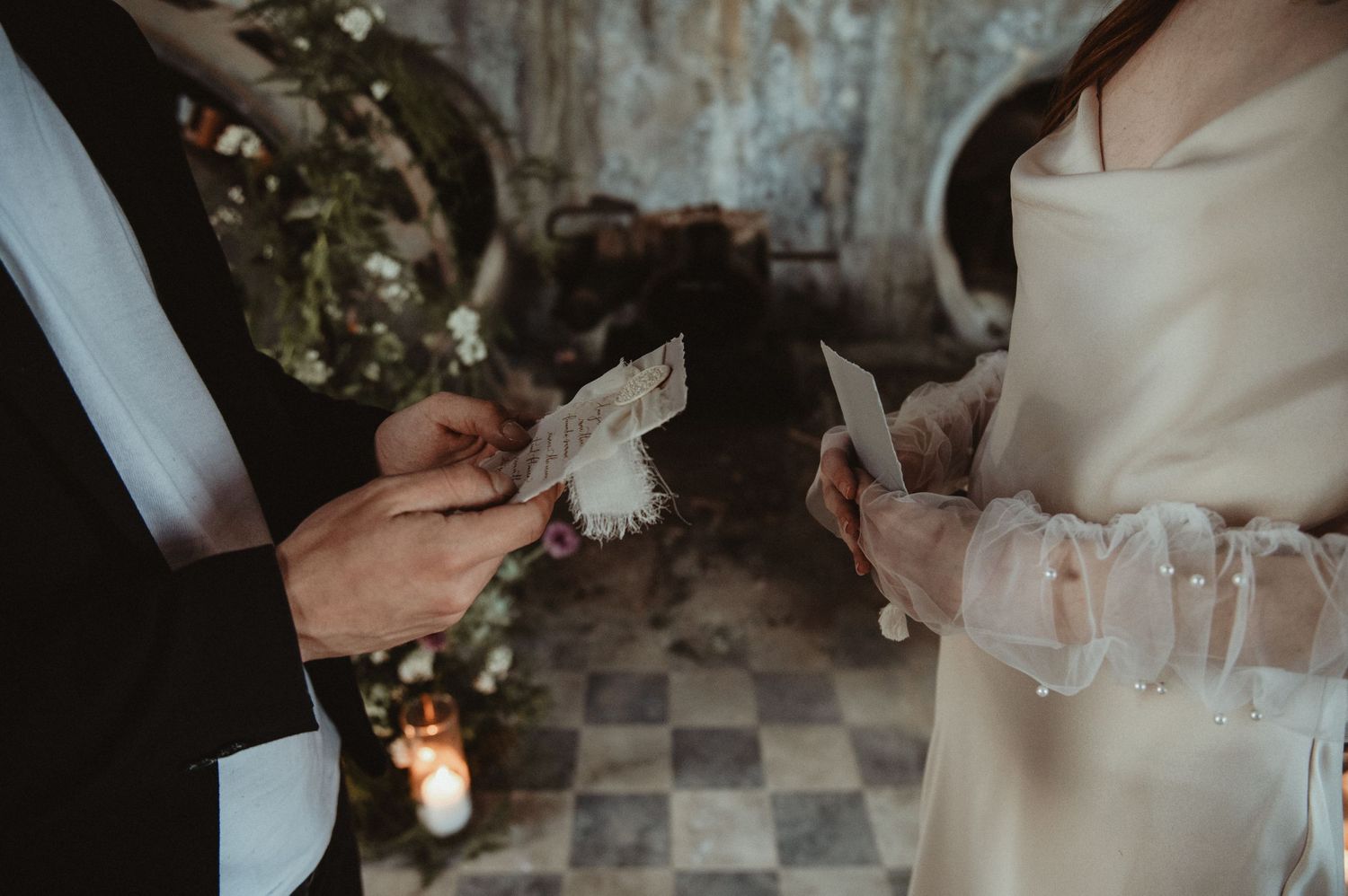 A couple exchanges vows while holding papers during an intimate ceremony in a rustic venue with a checkered floor.