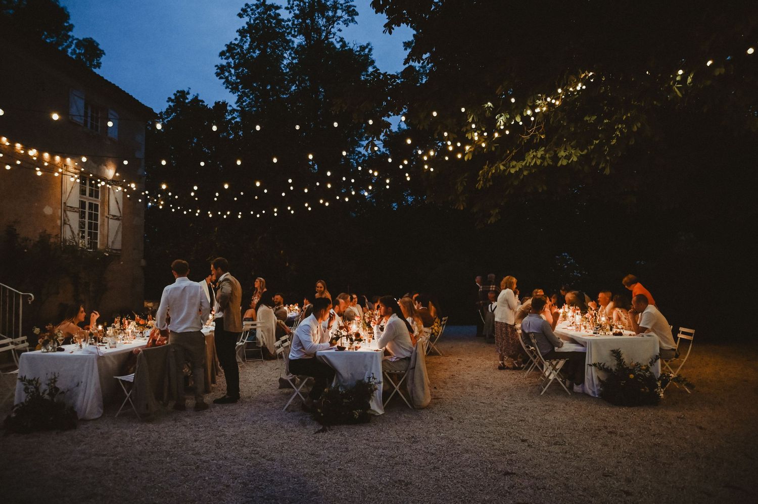 Evening wedding reception under string lights with guests seated at white-clothed tables.