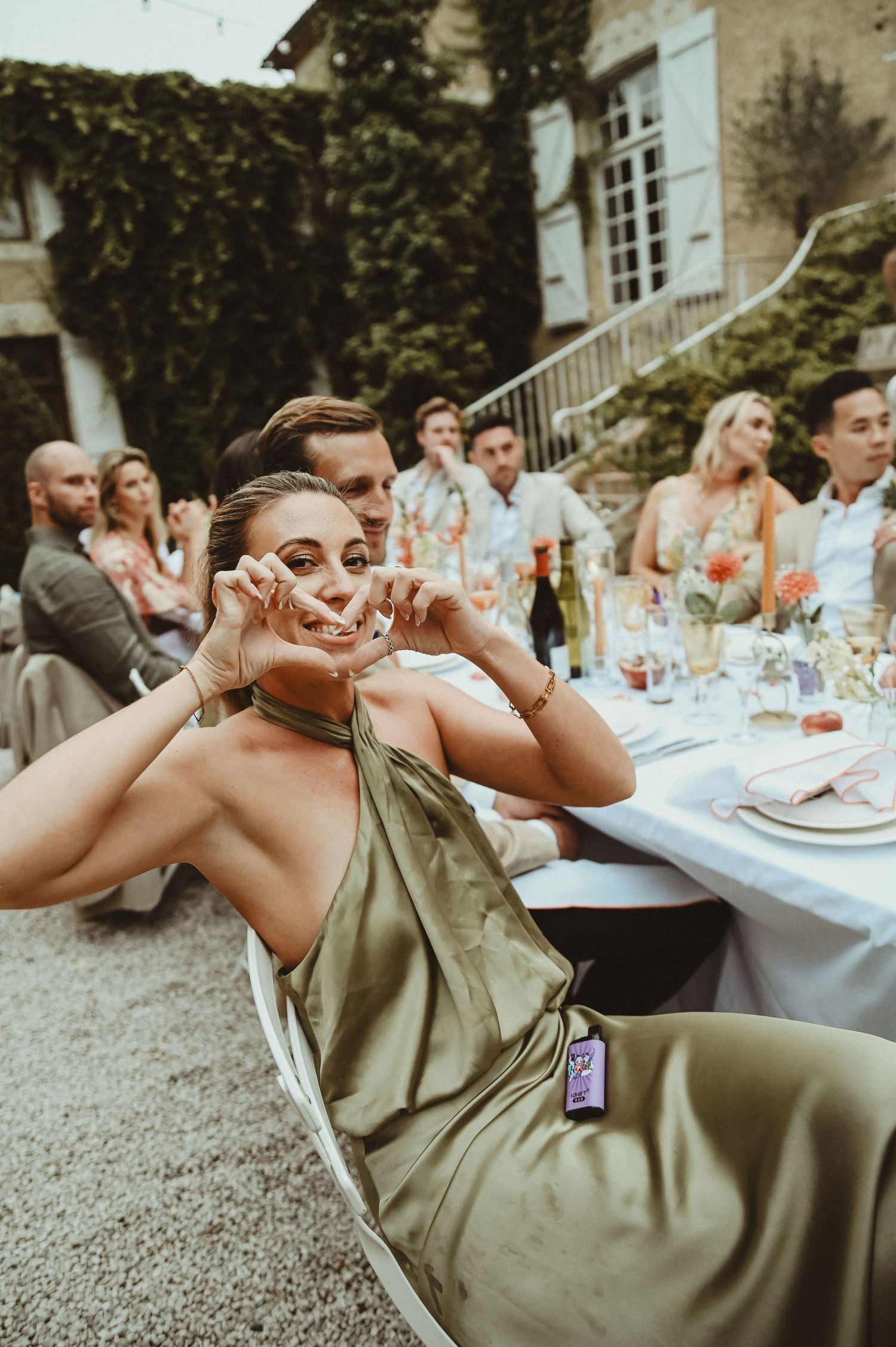 Wedding guest in olive green dress lounges at outdoor reception table in French countryside.