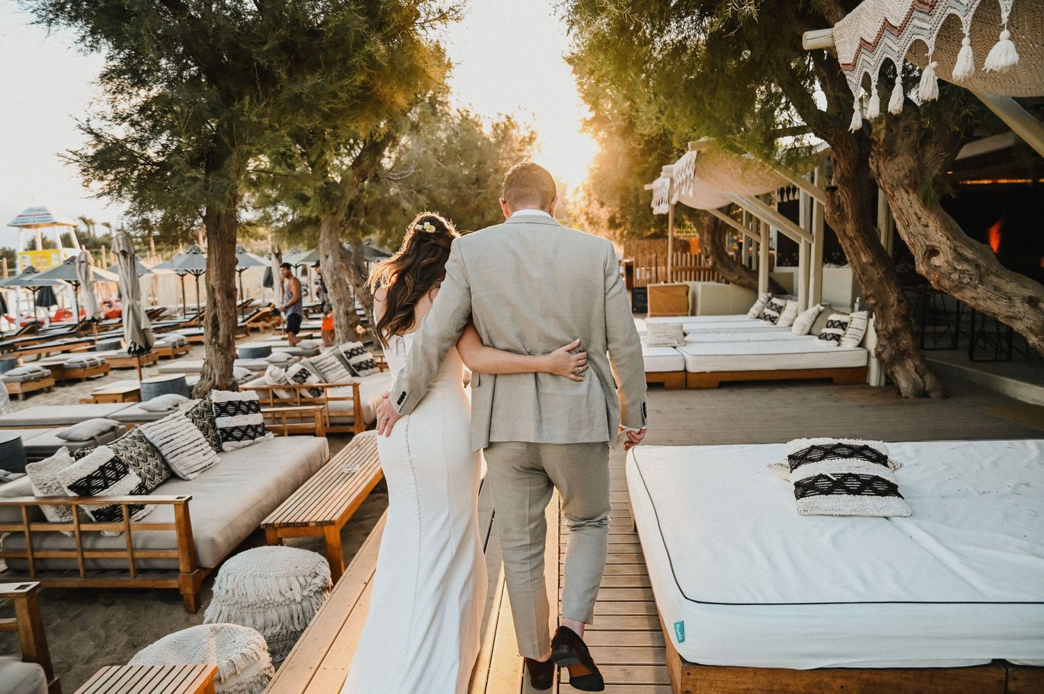 Newlyweds walking together at sunset among beach loungers and cabanas at a coastal venue.