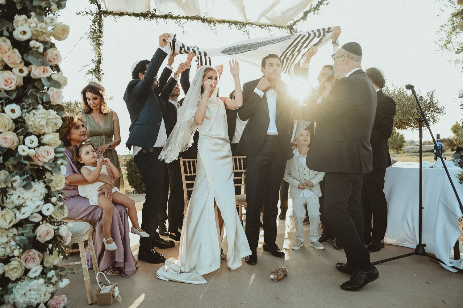 Wedding celebration under floral arch with guests cheering at sunset.