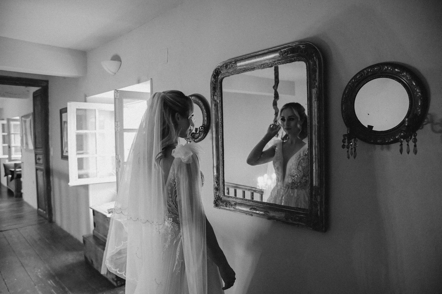 Black and white photo of a wedding dress being viewed in an ornate mirror in a hallway.