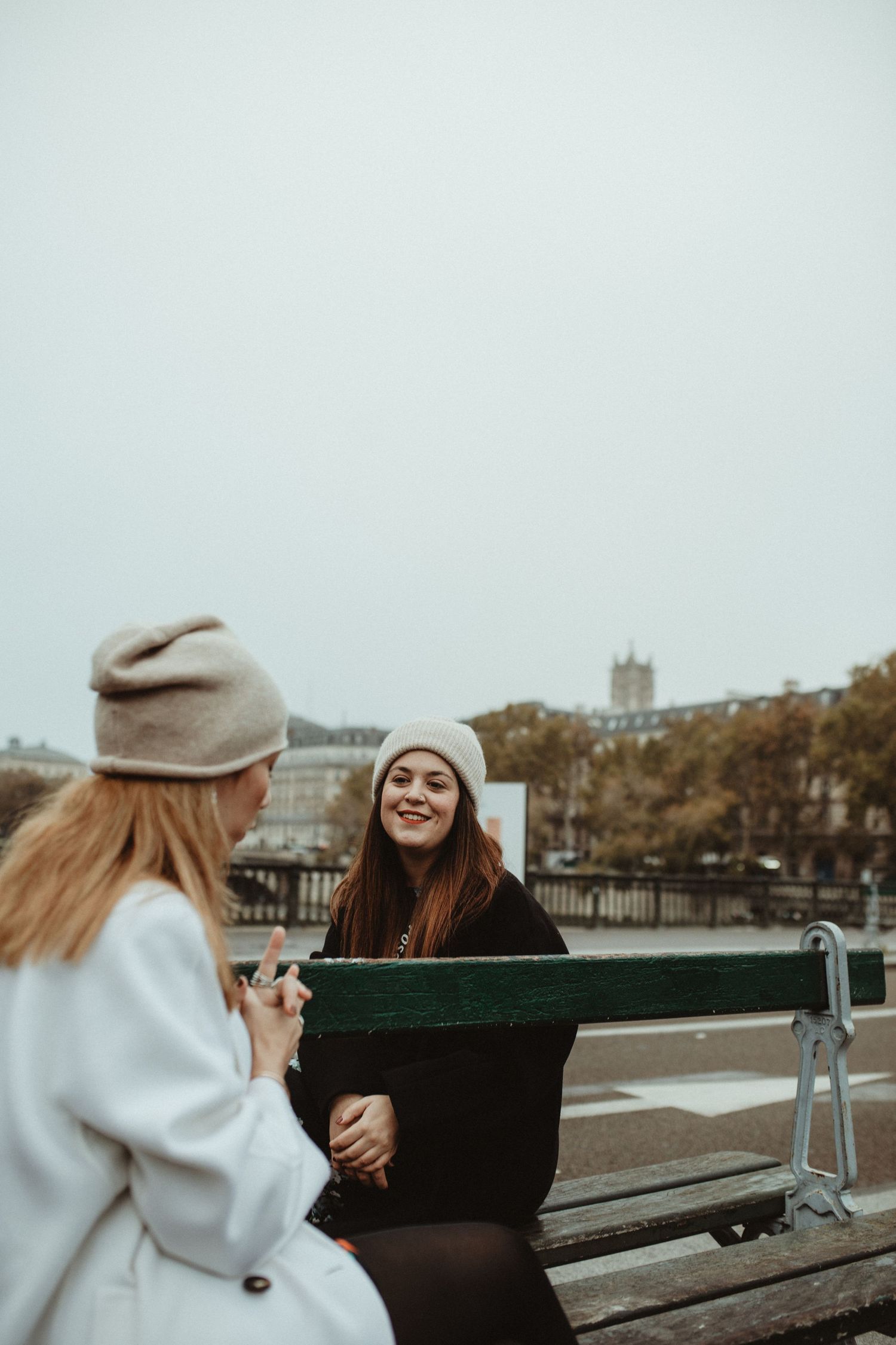 Two friends in warm winter clothing share a laugh while sitting on a bench on an overcast autumn day.