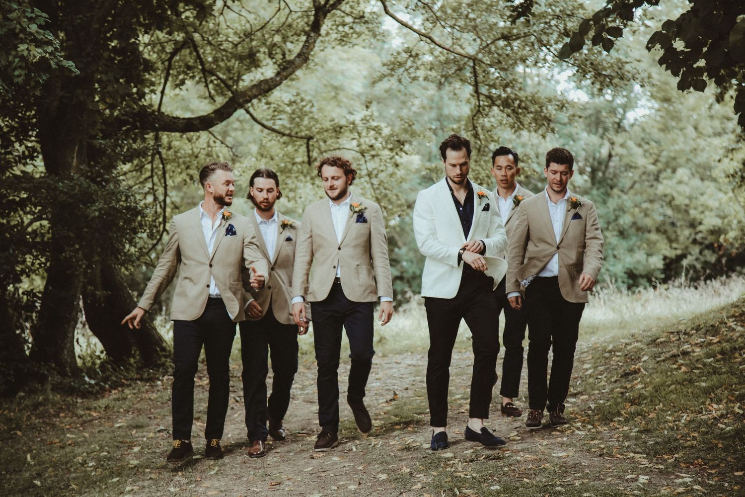 Groomsmen in light beige suits walking together through a wooded area.