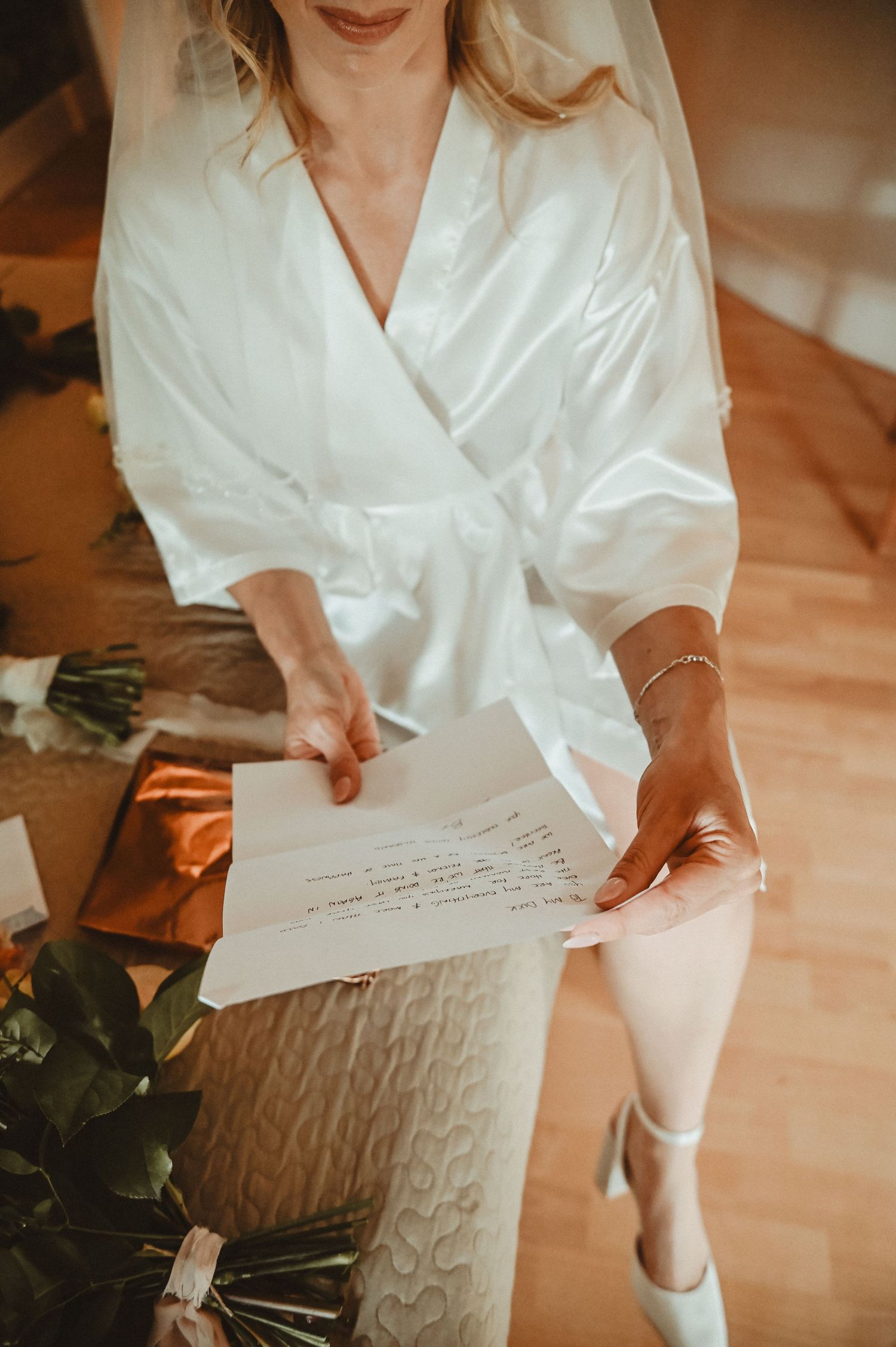 Person in white silk robe reading handwritten note among flowers and wedding preparations.